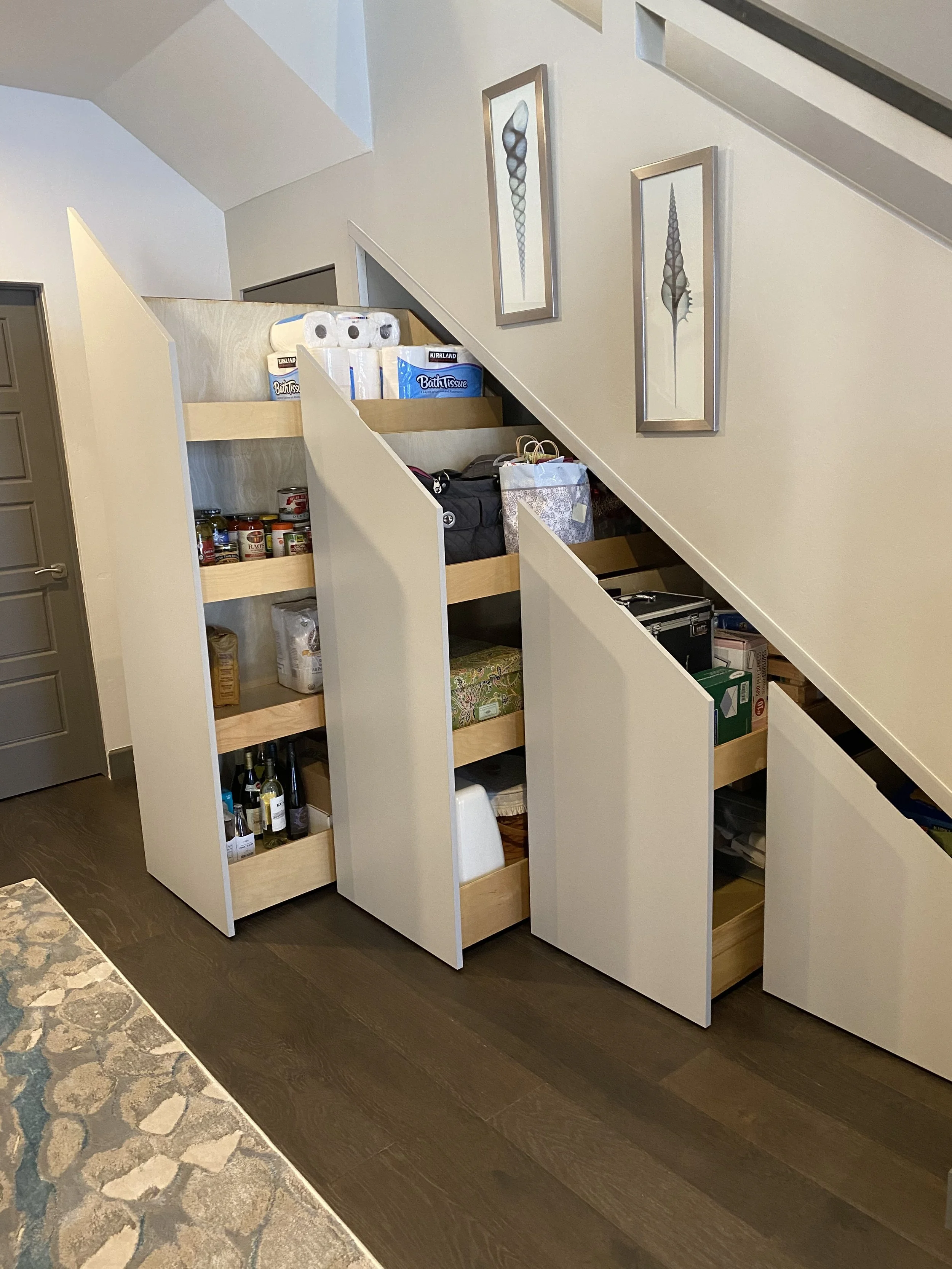 Interior of a house showing a partially assembled staircase storage area with shelves holding tissue paper, canned goods, and bottles, with framed artwork on the wall and a dark wood floor.
