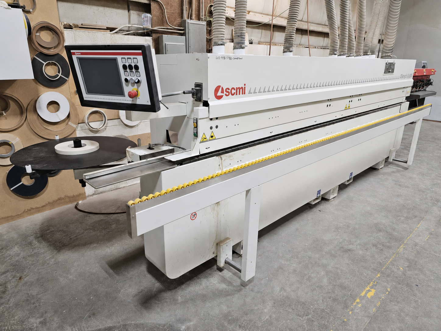 Industrial machine in a manufacturing facility, with a control panel and various rolls of material mounted on a wall nearby.