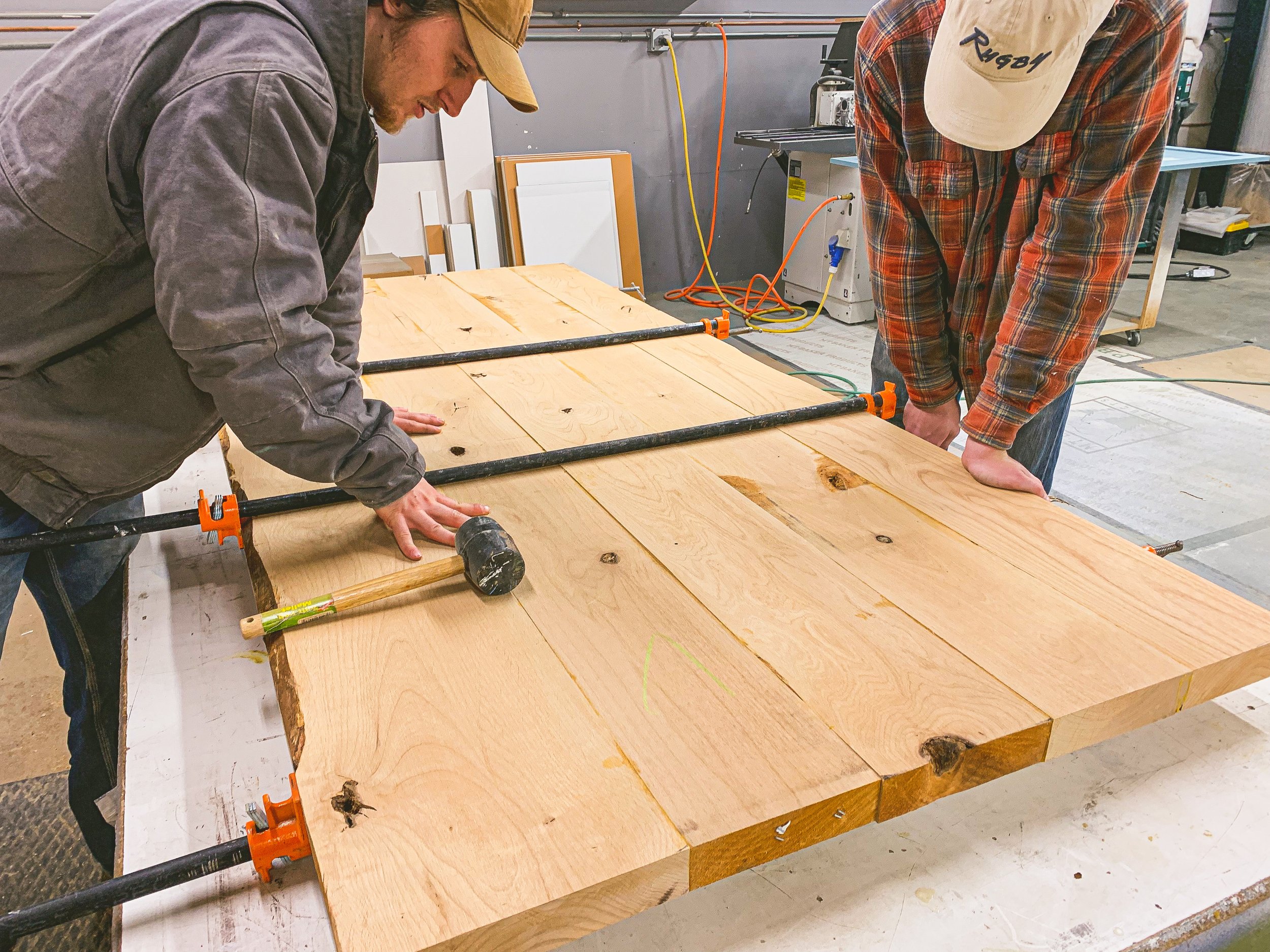Two people working on a wood project in a workshop, with wood clamps and large wooden slabs on a worktable.
