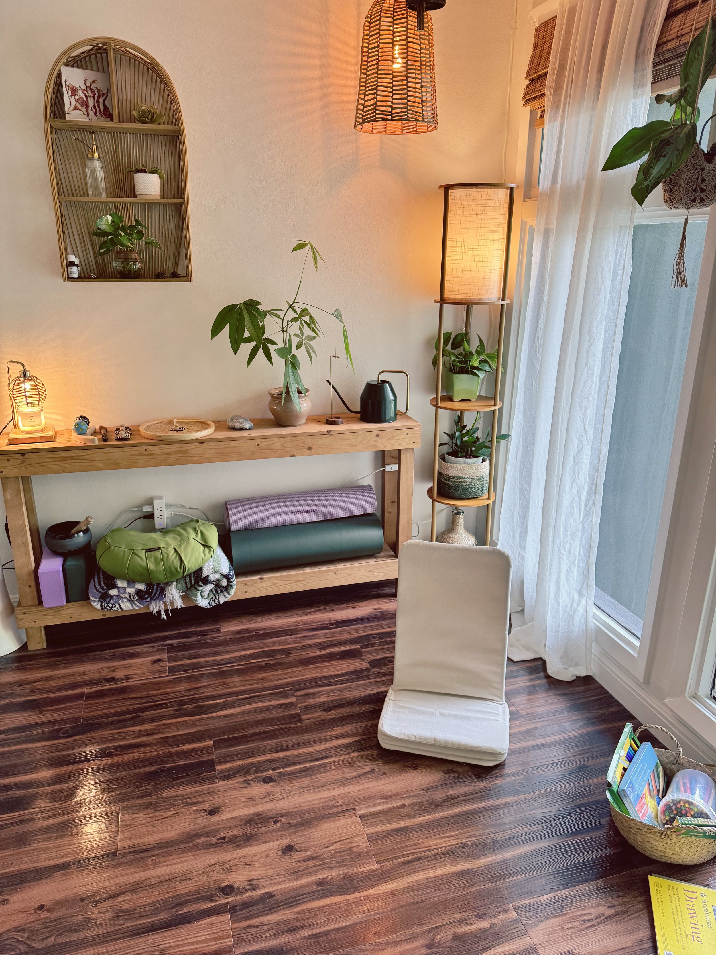 a floor chair next to a window, and a console table with plants and crystals on top and yoga mats and comfort items underneath