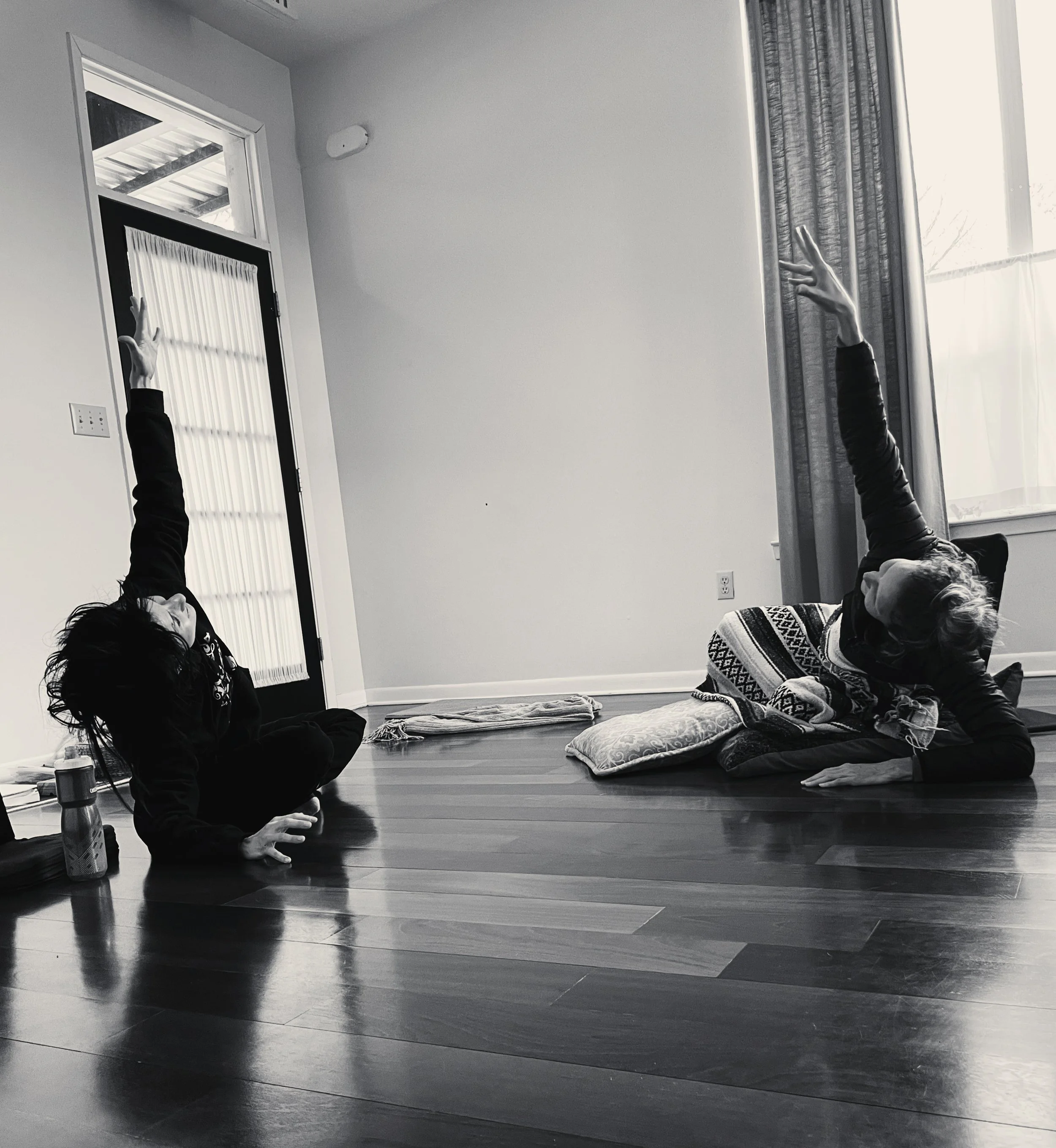 two people sitting across from each other on a studio floor; mirroring each other in movement; reaching towards the sky while leaning into the floor
