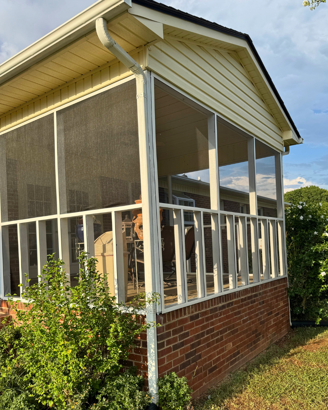 Enclosed porch with screened windows, brick foundation, and outdoor furniture, set against a partly cloudy sky.