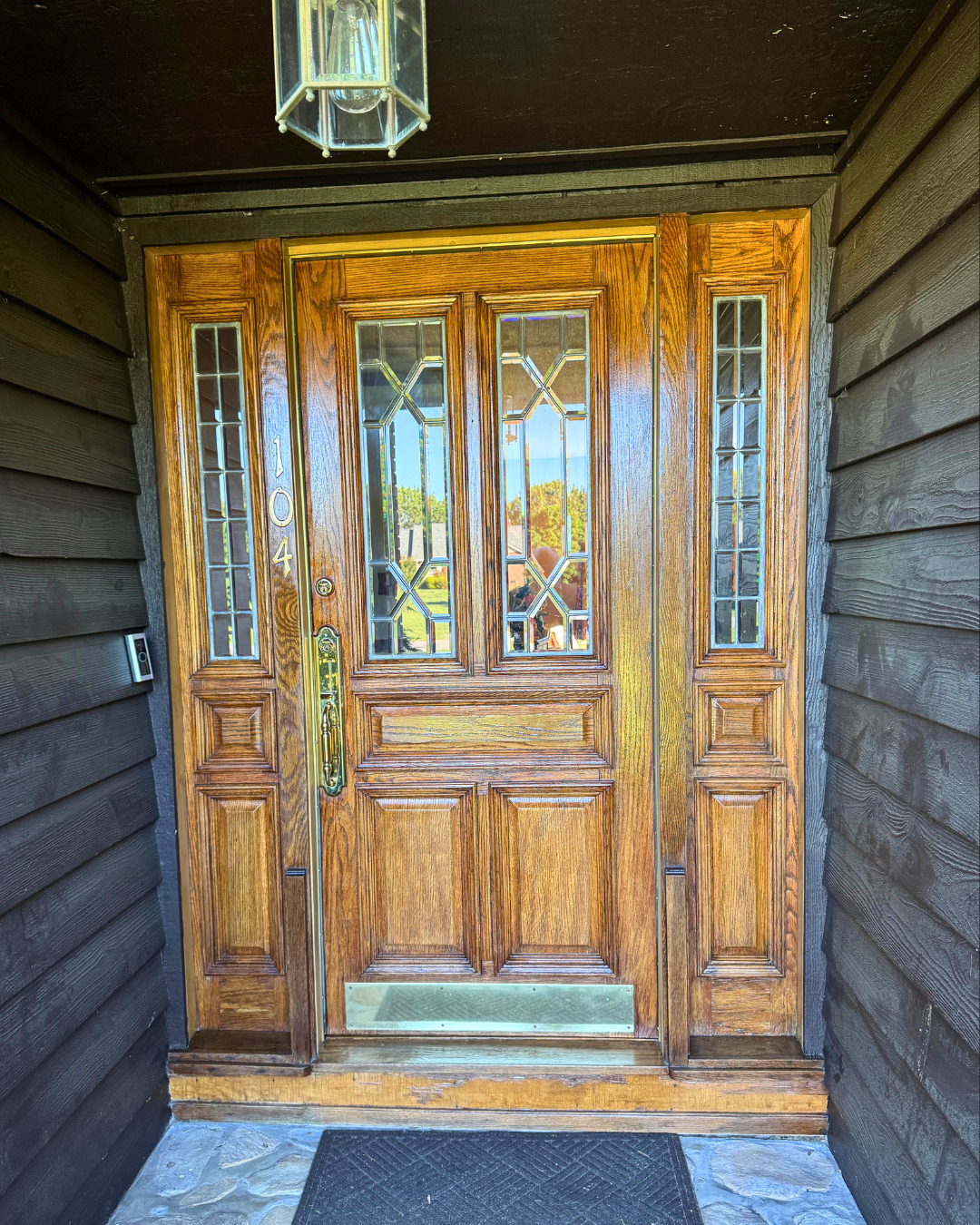 Wooden front door with decorative glass panels, house number 104 on the left, black doorbell on the left side, outdoor light fixture above, and black doormat on stone porch.