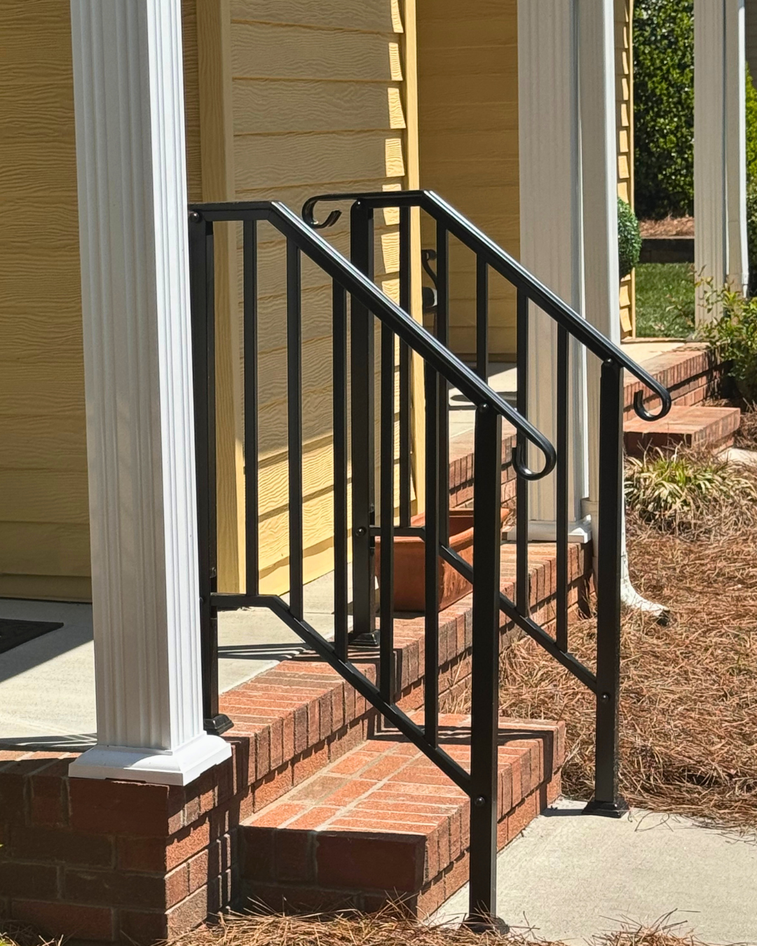 Black metal handrail on brick steps leading to porch of yellow house with white column and siding, surrounded by plants and trees in the yard.