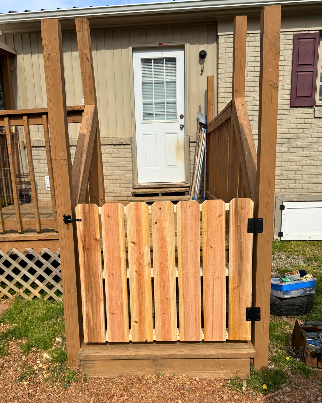 A partially built wooden deck and gate in front of a back door of a house with beige siding and brick. Building tools and supplies are nearby.