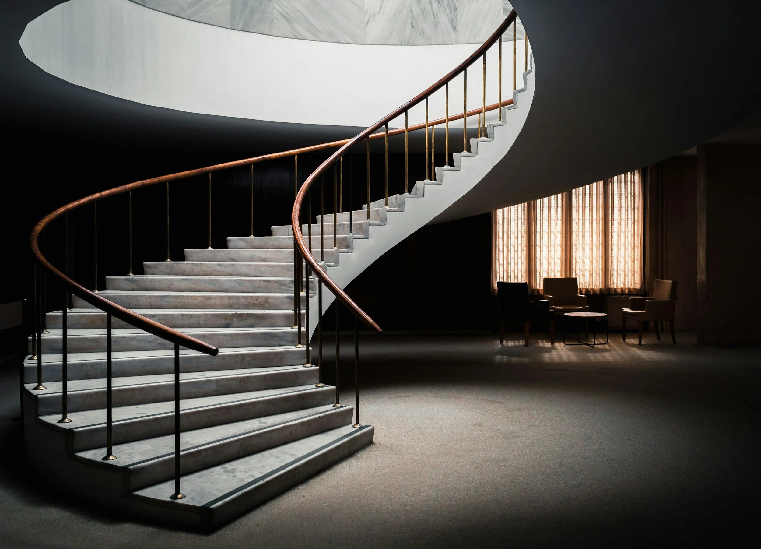 A staircase with white marble steps, a wooden handrail, and vertical golden balusters, inside a modern building with dark walls and large windows with vertical blinds.