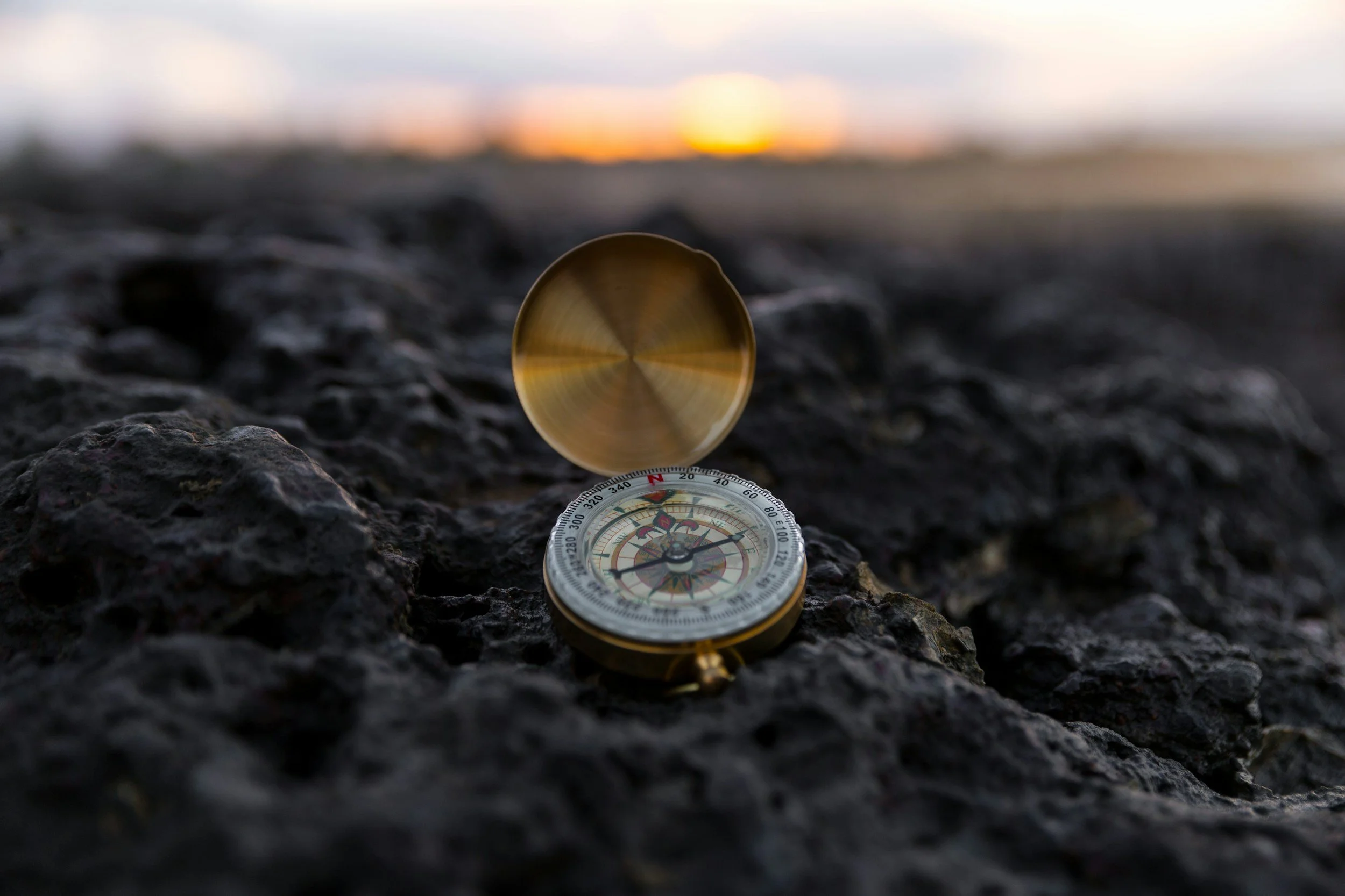 A compass lying on rocky ground at sunset, with the sun setting on the horizon and the sky partially cloudy.