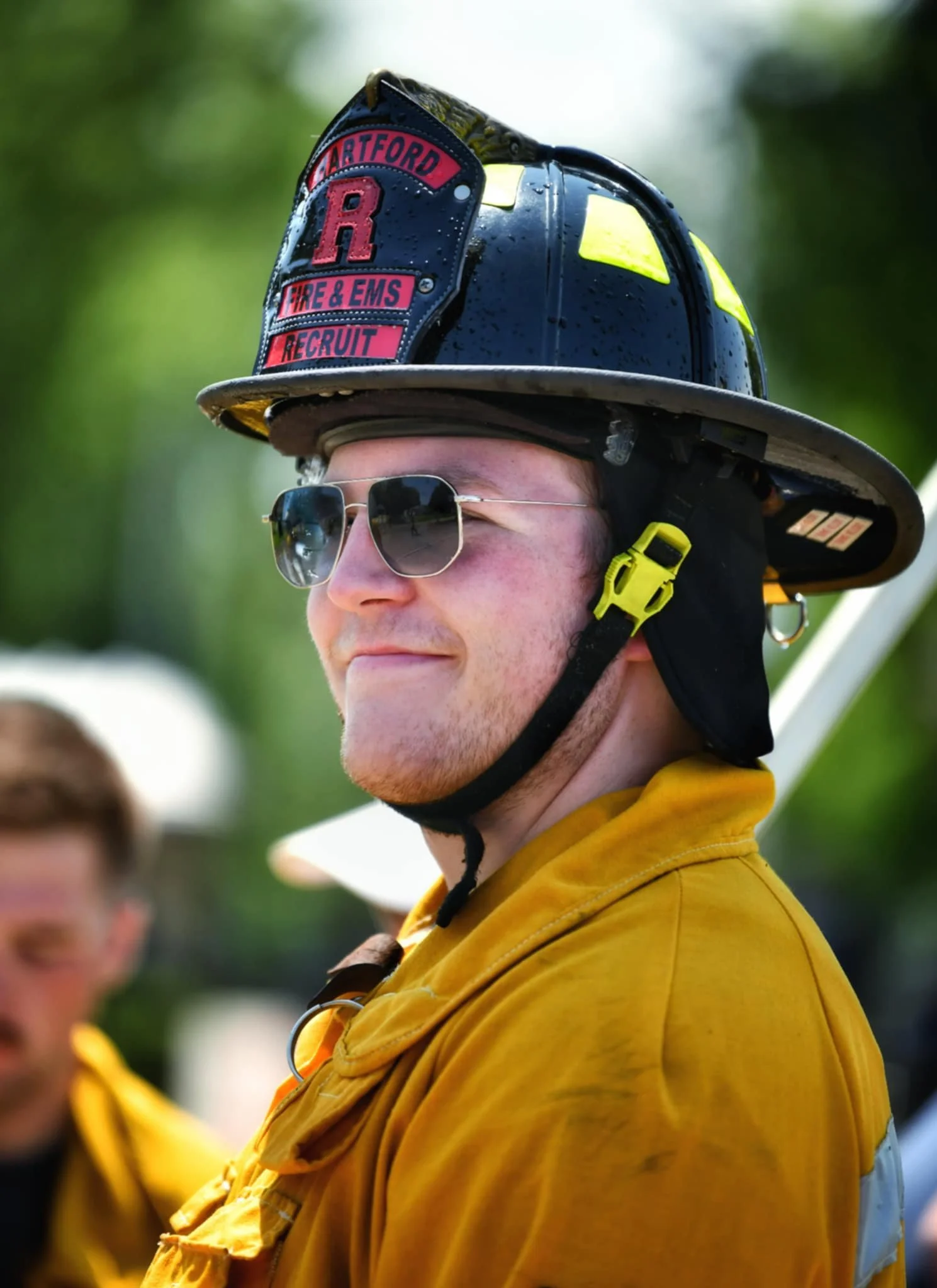 Firefighter wearing a black helmet with a tag reading 'Hartford Fire & EMS Recruit' and sunglasses, dressed in a yellow uniform, outdoors with blurred green trees in the background.