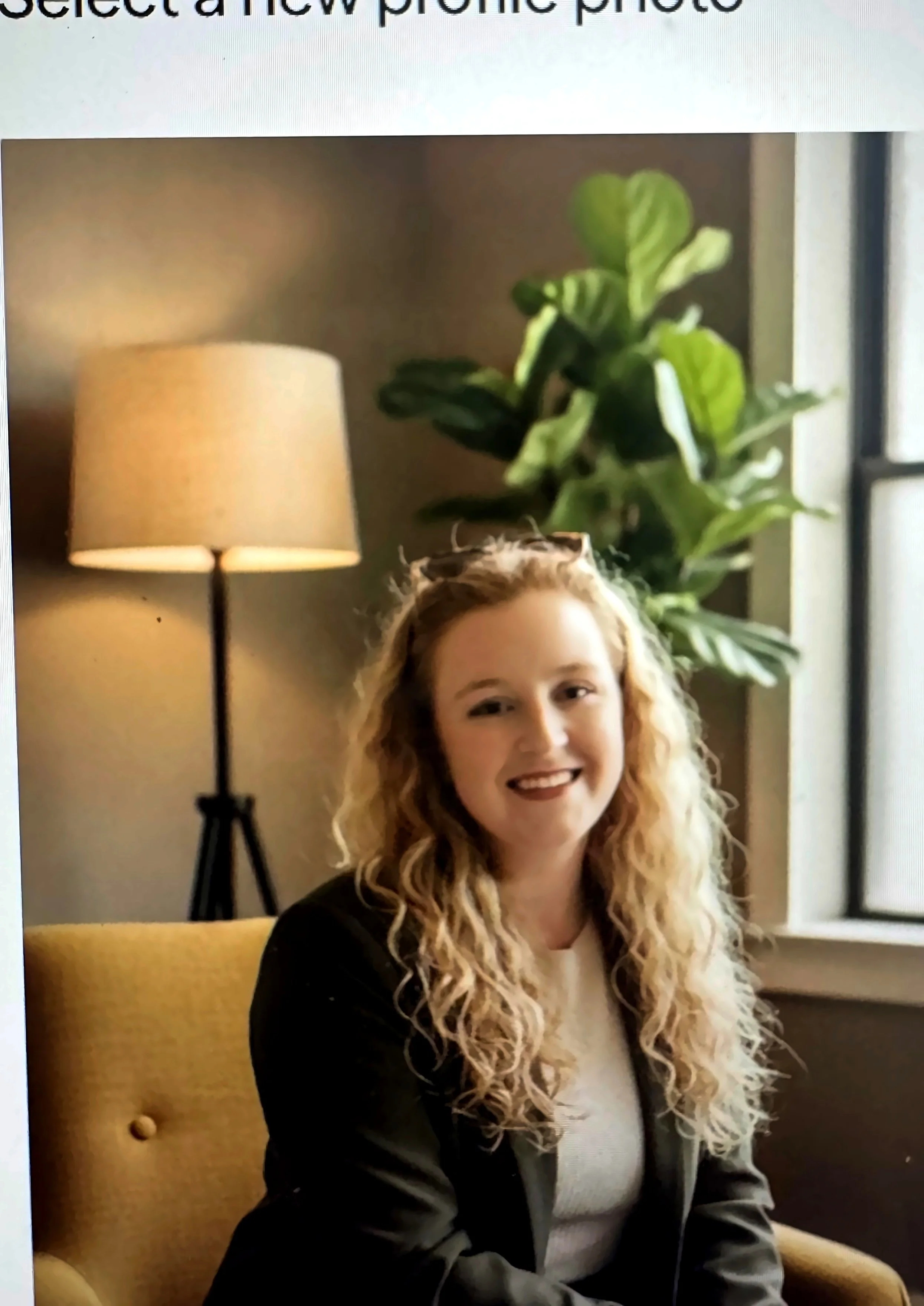 A woman with a welcoming smile, curly strawberry blonde hair sitting on a yellow chair in front of a green houseplant, with a beige lamp with a lampshade in the background.