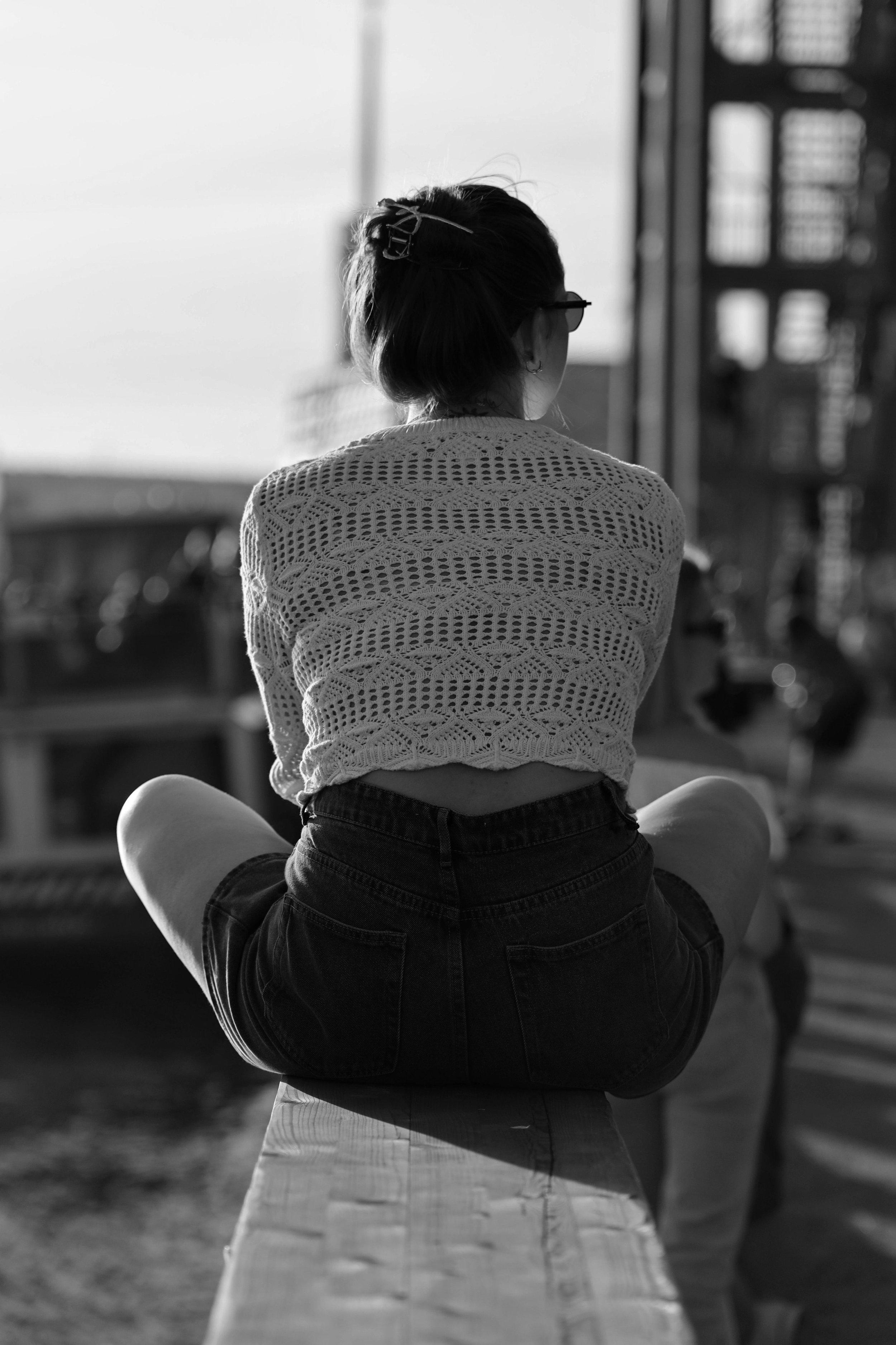 Black and white image of a woman sitting alone on a ledge, back turned, evoking isolation and quiet unease.