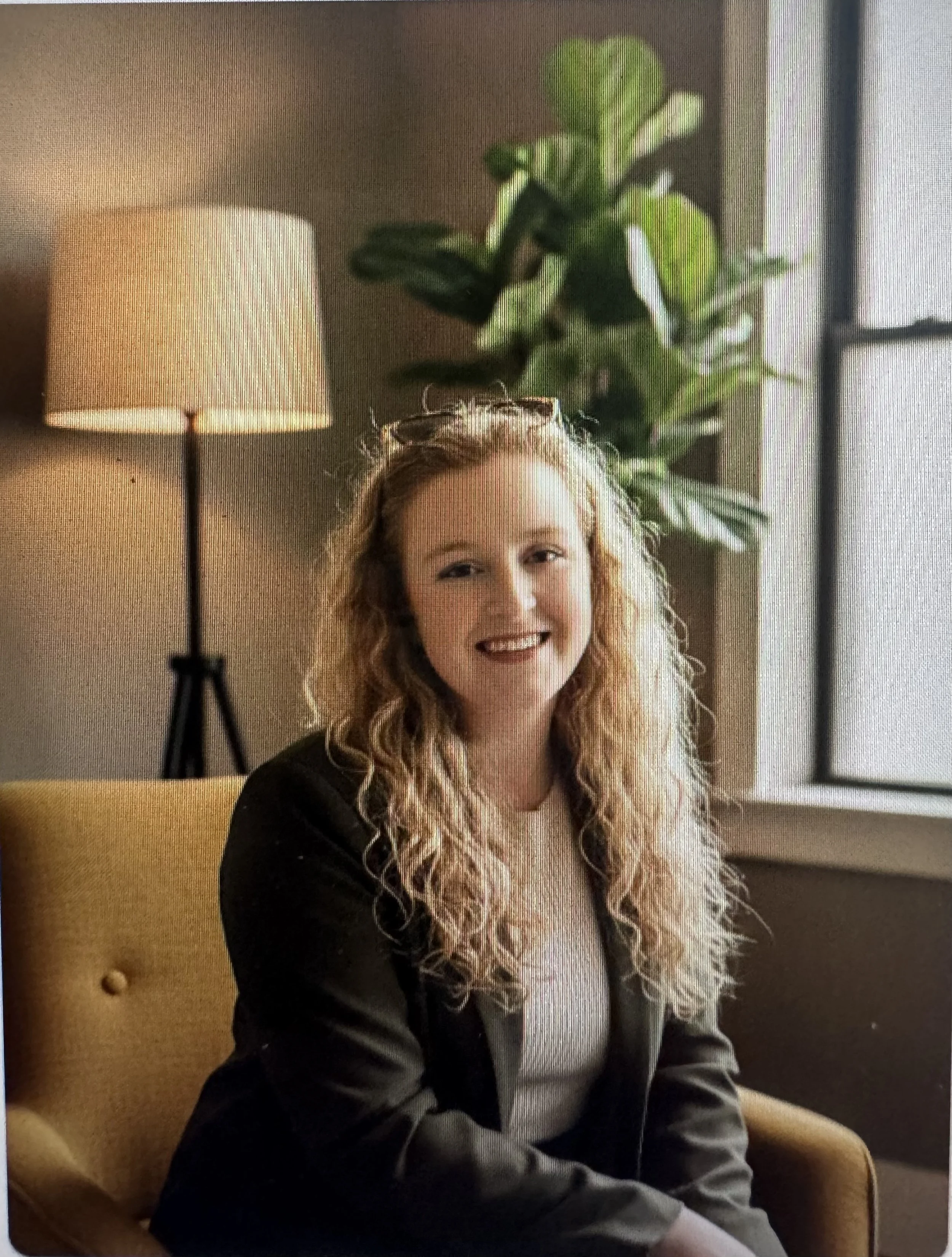 Founder, Chloe Melka, with curly strawberry-blonde hair sitting in her office on a yellow chair, smiling. Behind her is a tall green plant, a lamp, and a window with a white curtain.