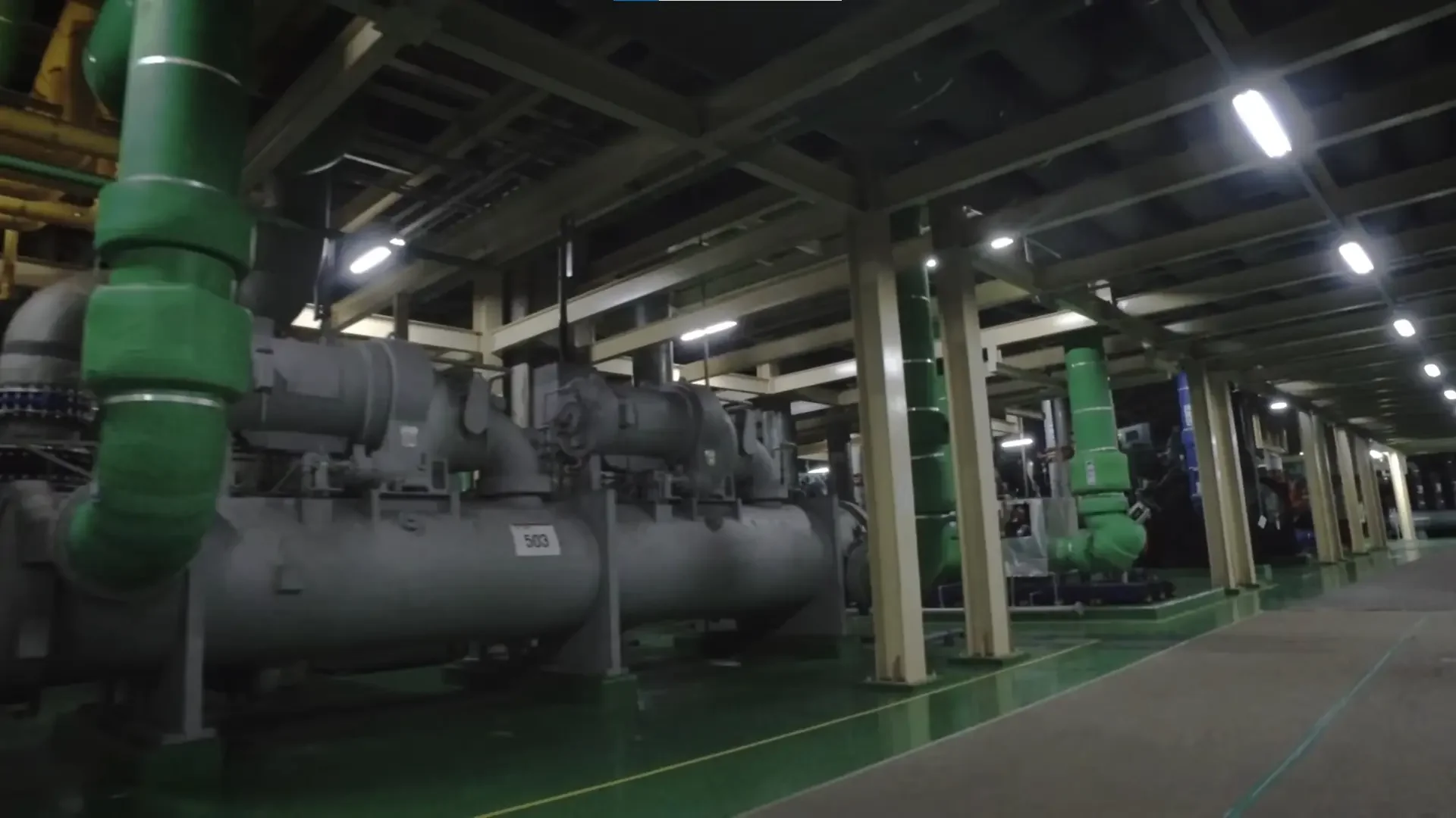 Imagen de una sala de máquinas industrial con tuberías grandes y equipos mecánicos, iluminación artificial y estructura metálica en el techo.