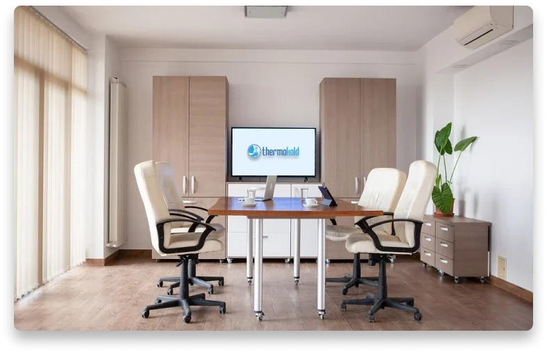 Sala de reuniones con mesa de madera, cuatro sillas blancas con brazos, un monitor con el logo de ThermoFrost, un portátil y teléfonos en la mesa, armarios de madera eléctrica y una planta en una mesa auxiliar.