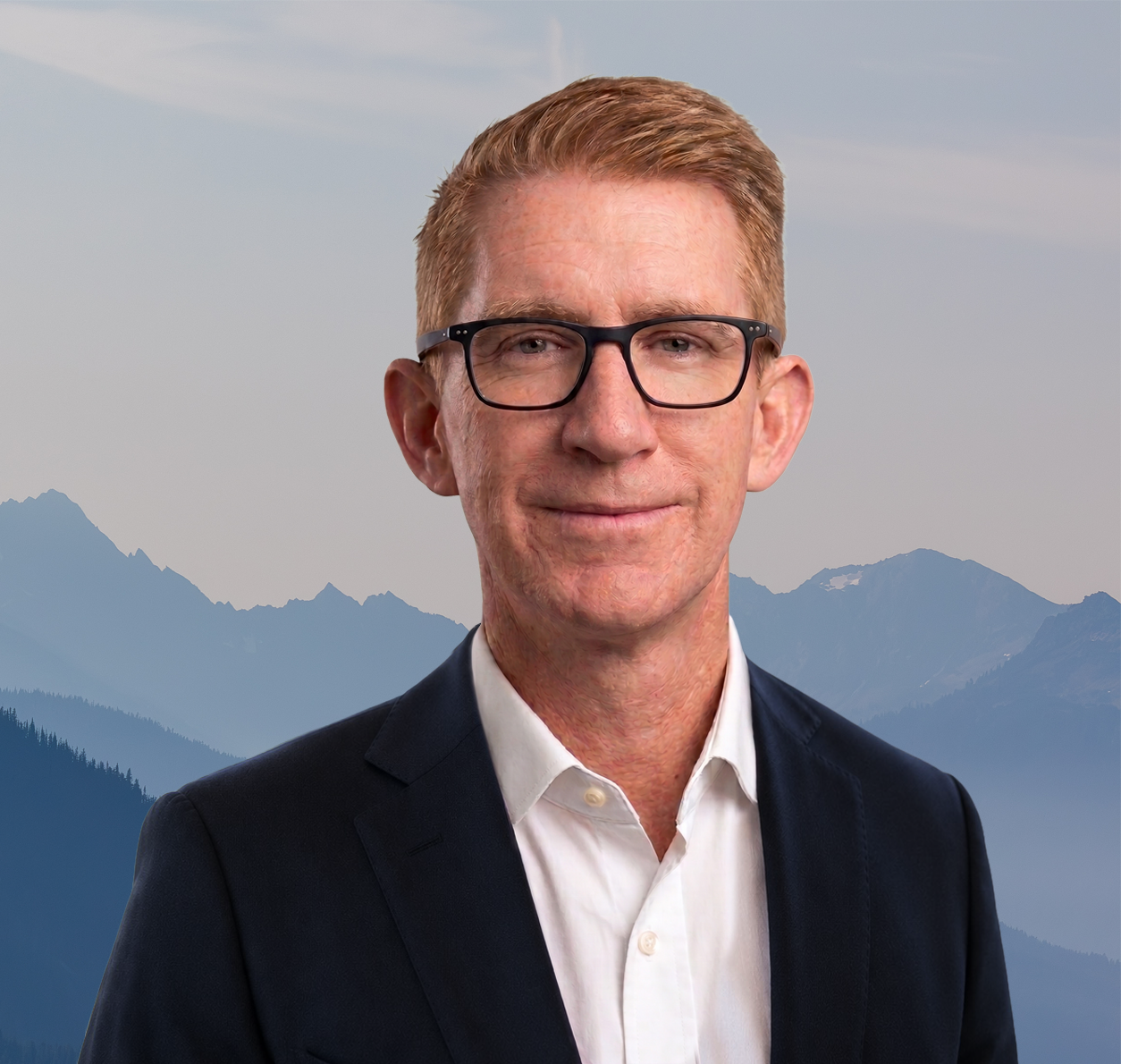 Headshot of a man with glasses, wearing a suit and white shirt, with mountains in the background.