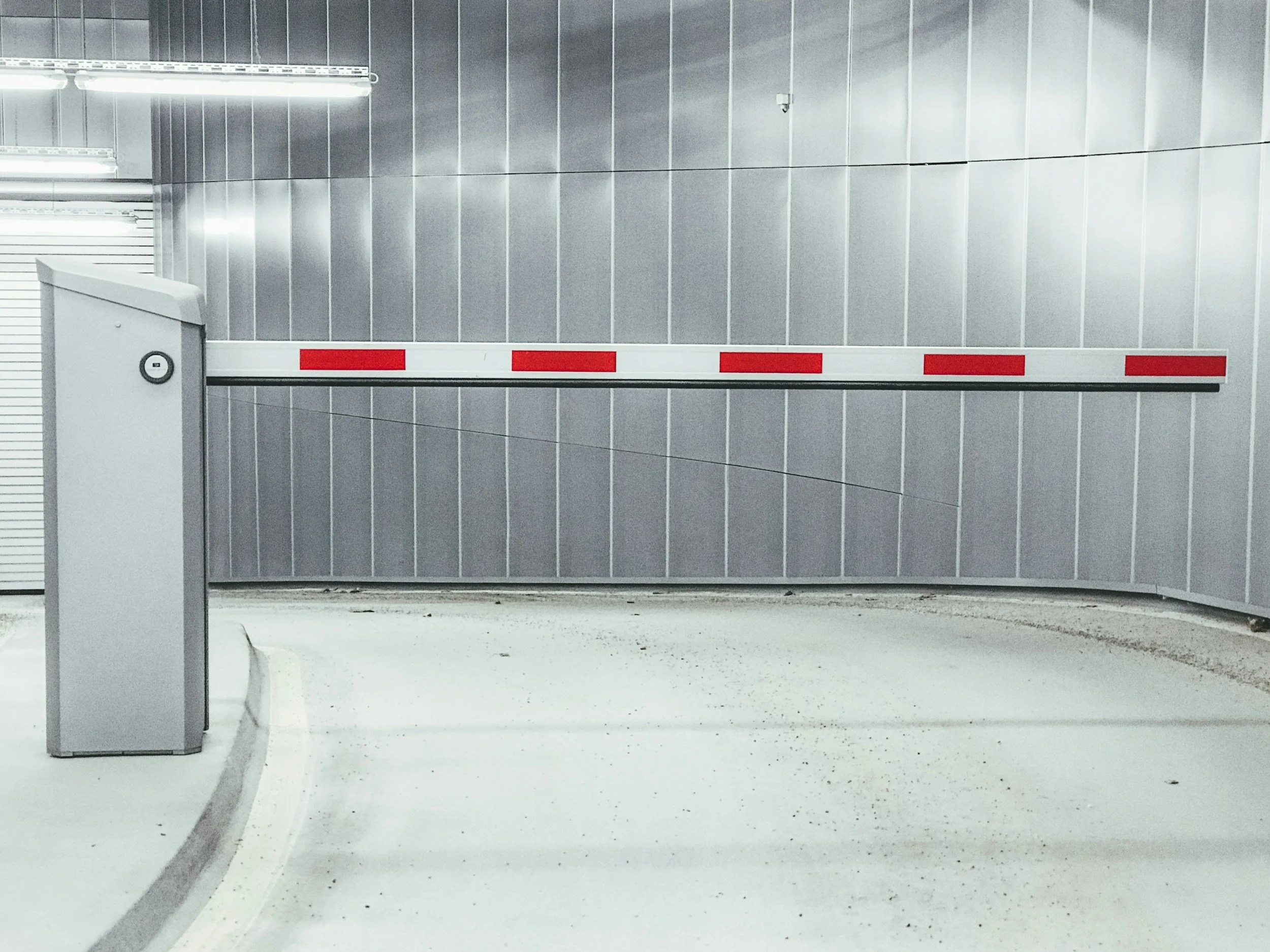 Una barrera de entrada en una estacionamiento. La barrera es blanca con franjas rojas y hay una estructura en color gris a la izquierda.