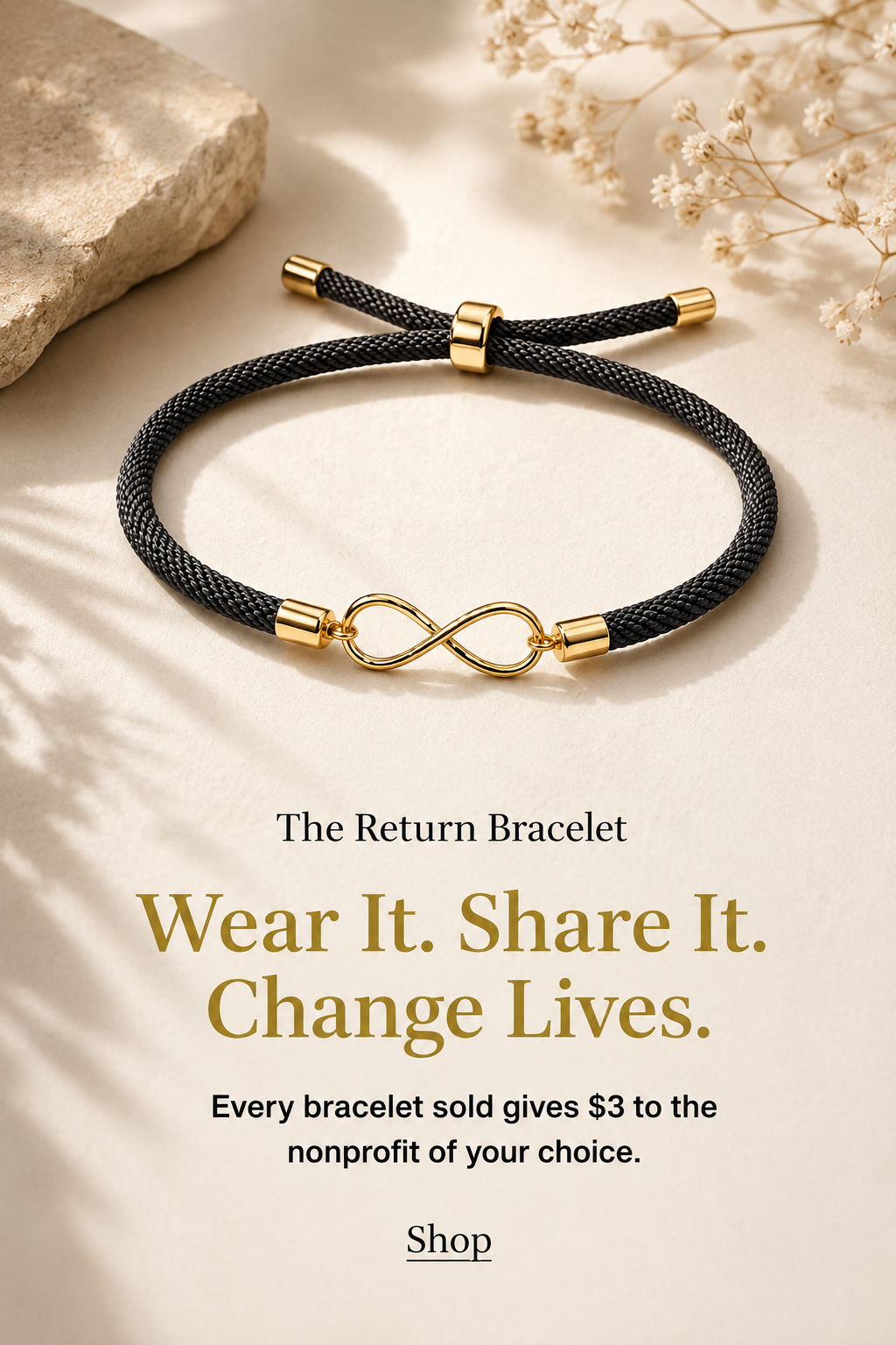 A black braided bracelet with a gold infinity symbol in the center, gold caps at each end, and a gold clasp, placed on a beige surface with a stone to the left and white flowers to the right.