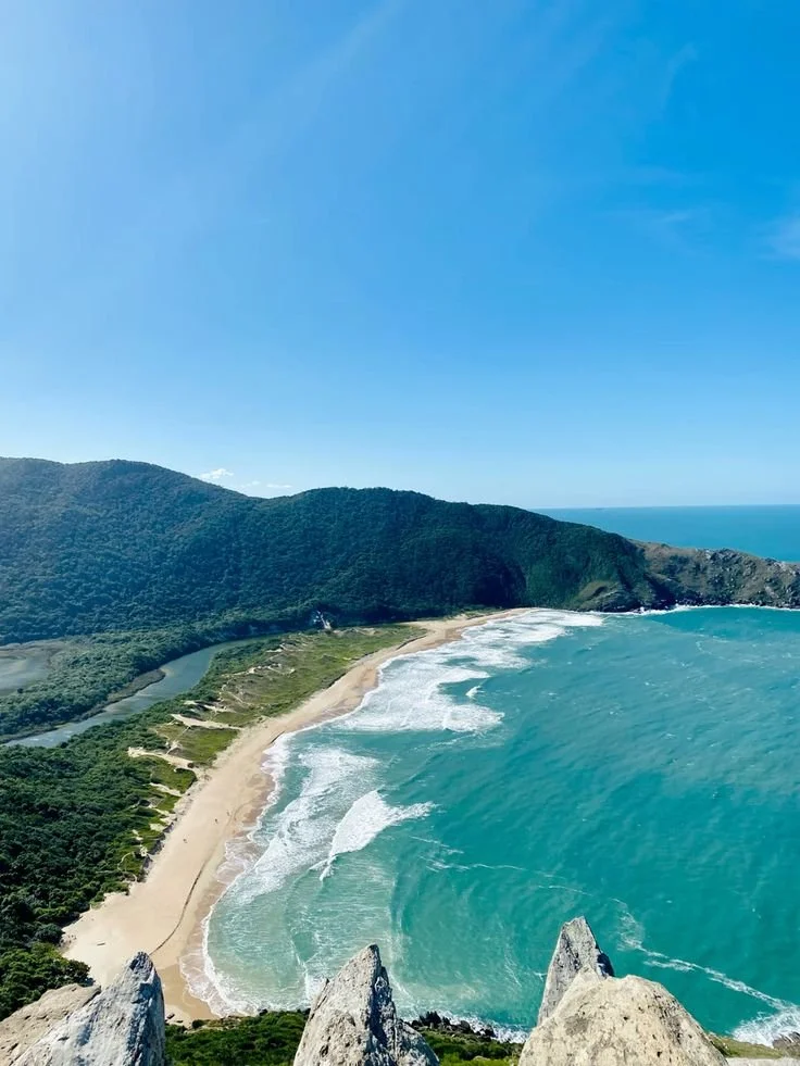 Vista aérea de una playa con arena dorada y olas rompiendo, rodeada de montañas verdes y un cielo despejado