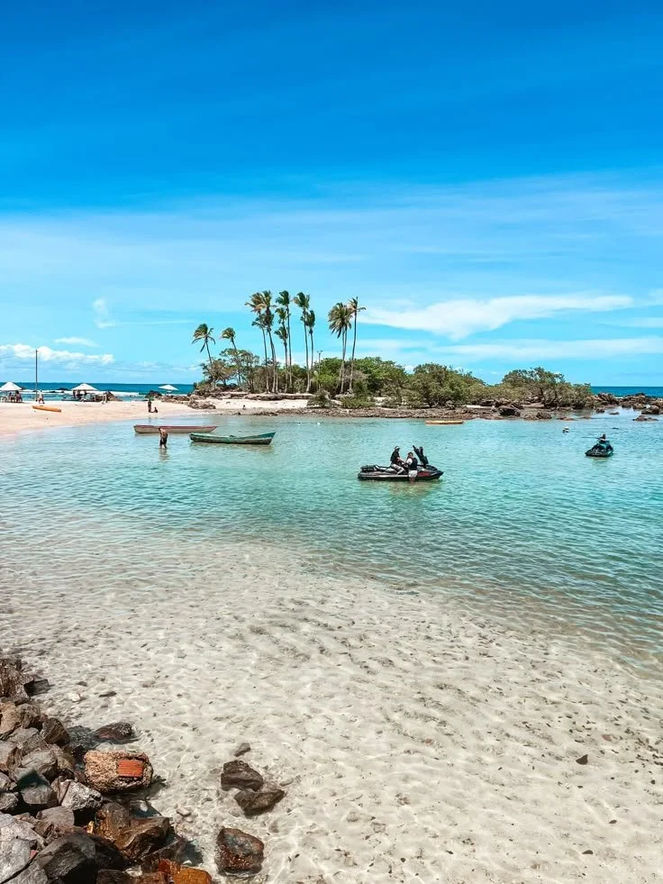 Playa tropical con aguas claras y palmeras en una pequeña isla, con personas en botes y en la playa, cielo azul y pocos nubes.