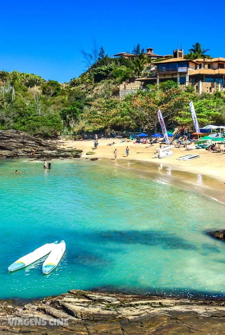 Playa con arena dorada, agua turquesa, personas nadando, palmeras y casas en colina con cielo azul claro.
