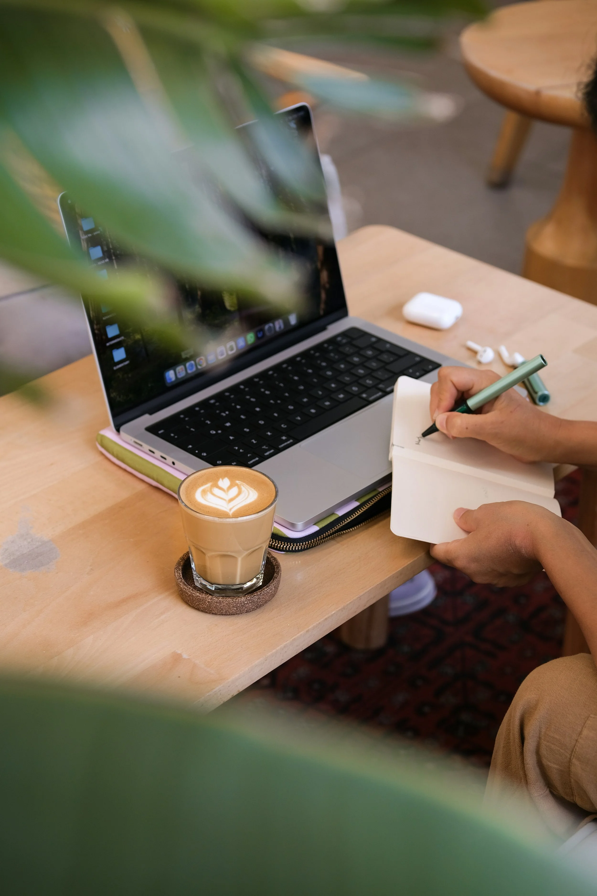 A person writing in a small notebook at a wooden table with a laptop, a glass of coffee with latte art, and wireless earbuds on the table, with some green leaves partially covering the view.