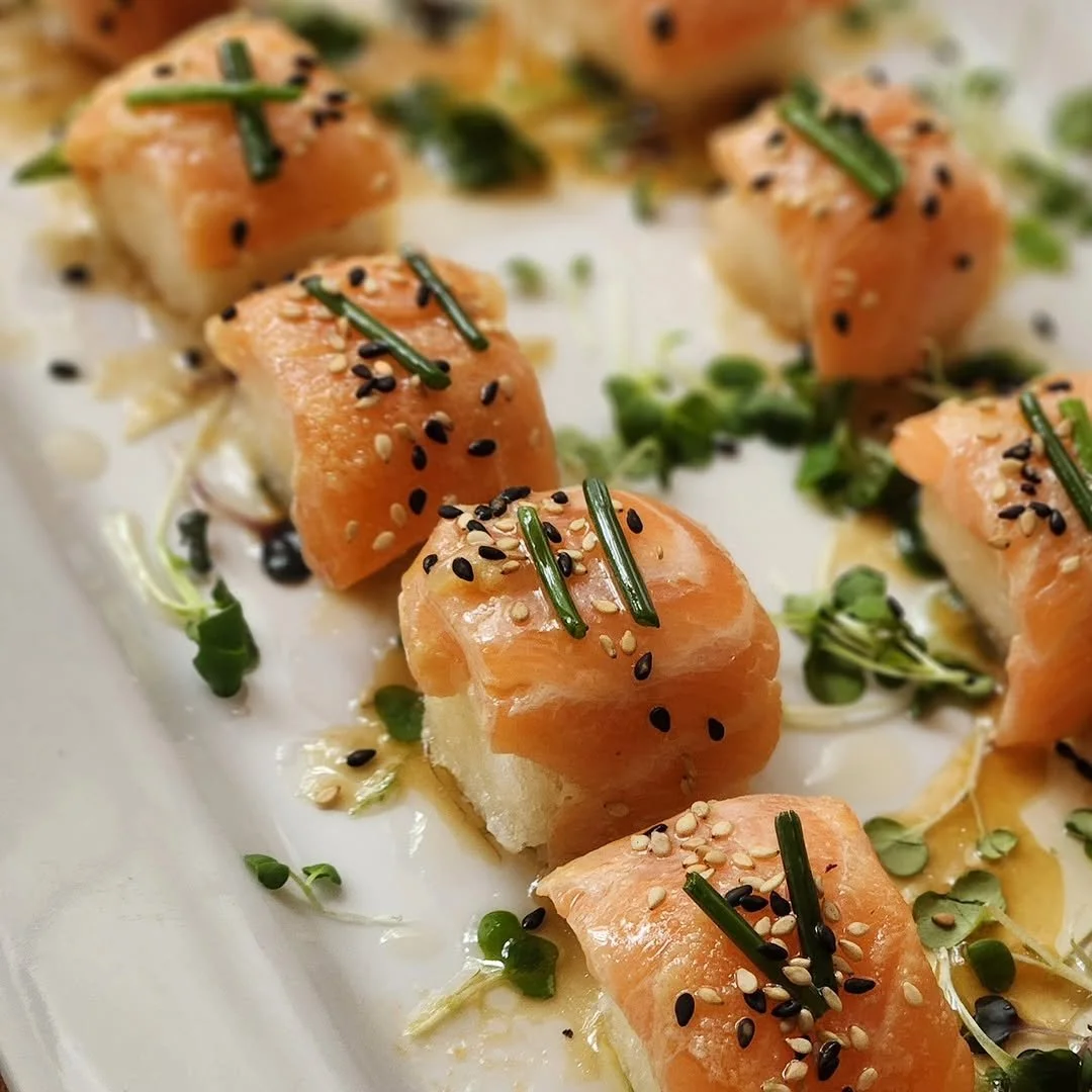 Close-up of bite-sized pieces of sushi topped with salmon, sesame seeds, chives, and microgreens on a white platter