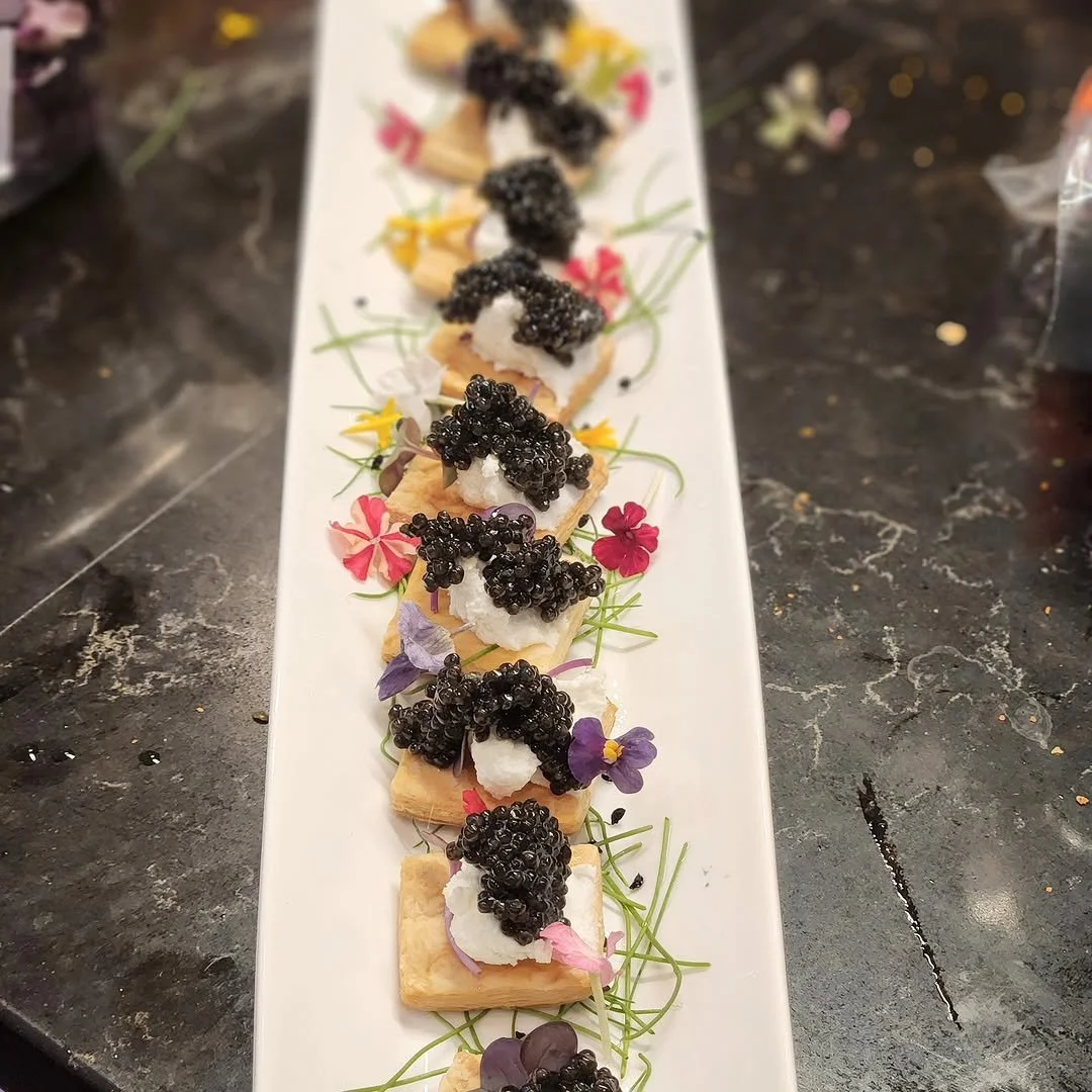 Appetizer of crackers topped with cheese, caviar, and edible flowers, arranged on a long white platter garnished with microgreens.