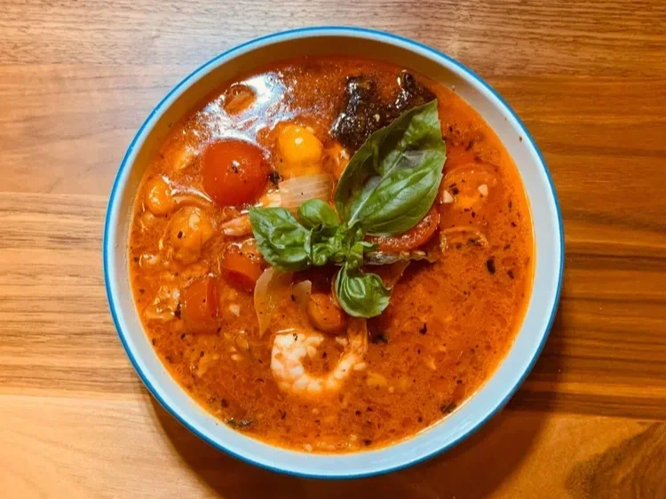 Bowl of spicy tomato-based seafood soup garnished with basil, cherry tomatoes, and herbs on wooden table
