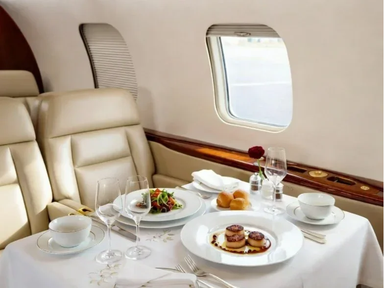 Luxury airplane first-class dining table with salad, bread rolls, dessert, white tablecloth, and glassware inside an aircraft with beige leather seats and windows.