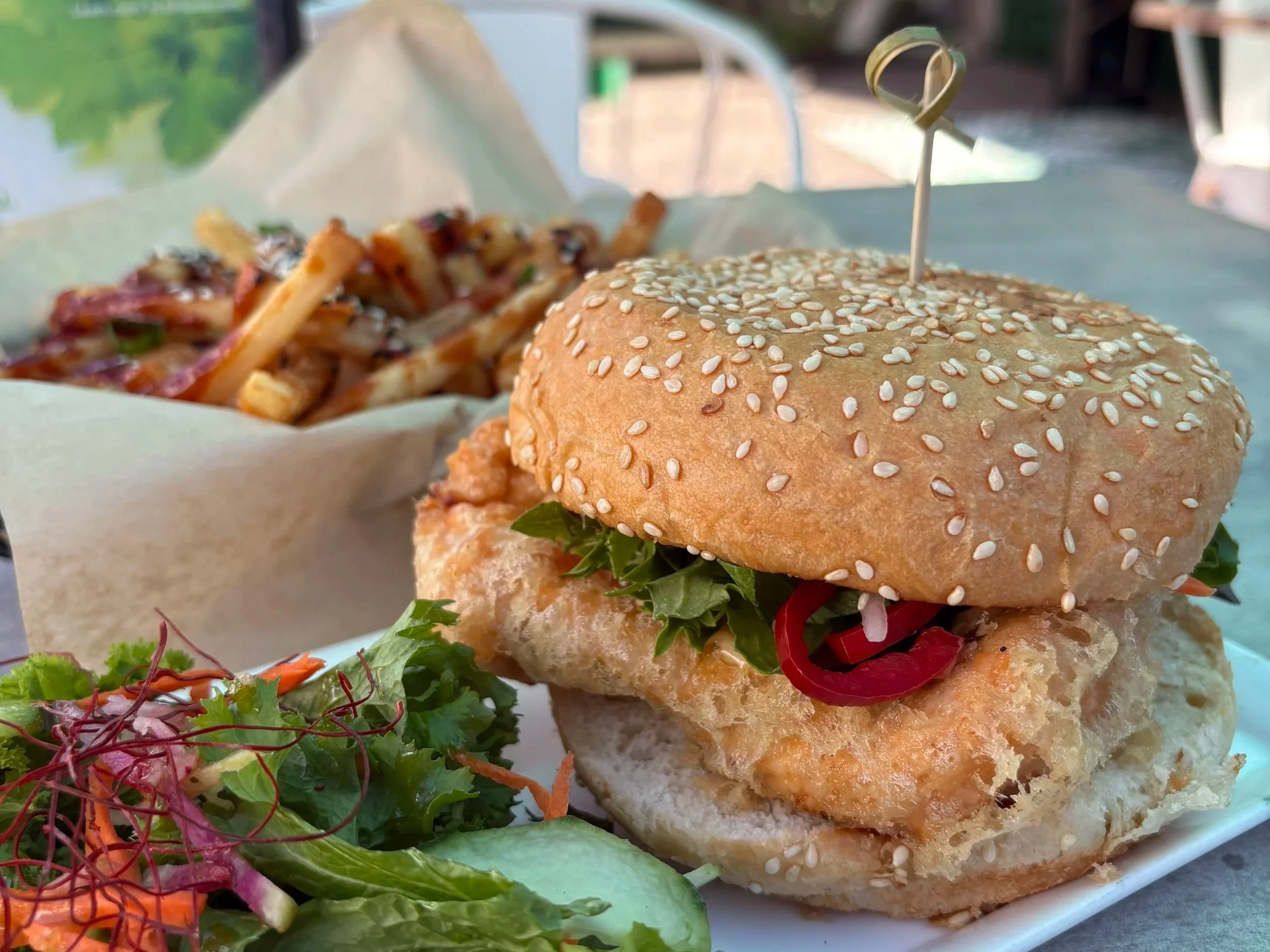 Close-up of a sesame seed bun sandwich with lettuce and tomato, served with a side of mixed greens and shredded carrots.