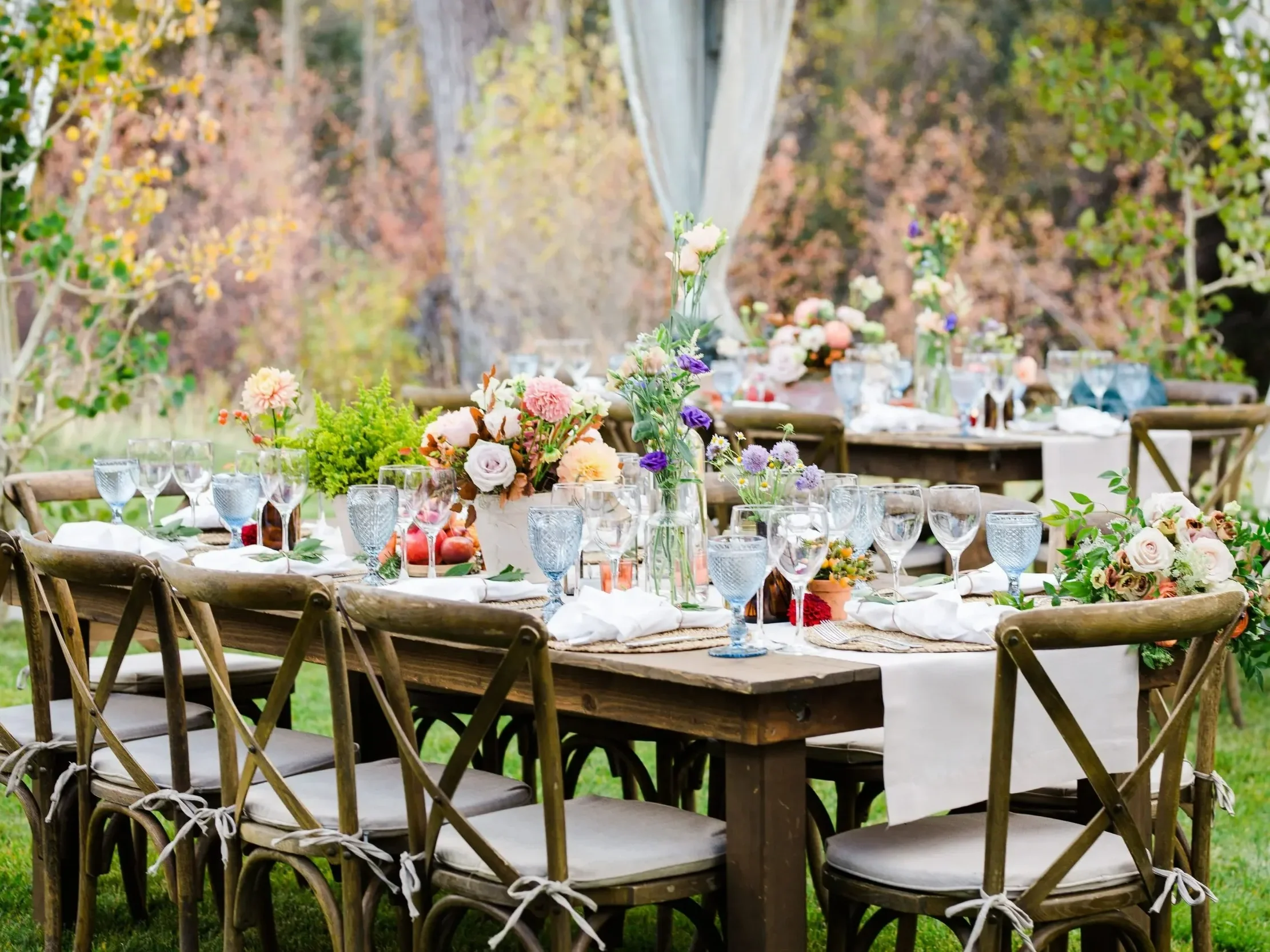 A long wooden table decorated with colorful flowers in vases and surrounded by wooden chairs with white cushions, set outdoors in a lush green garden with trees in the background, for a celebration or event.