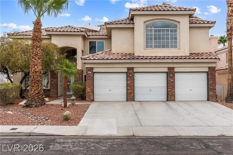 A two-story suburban house with a three-car garage, beige stucco exterior, red brick accents, tiled roof, and desert landscaping including palm trees and bushes.