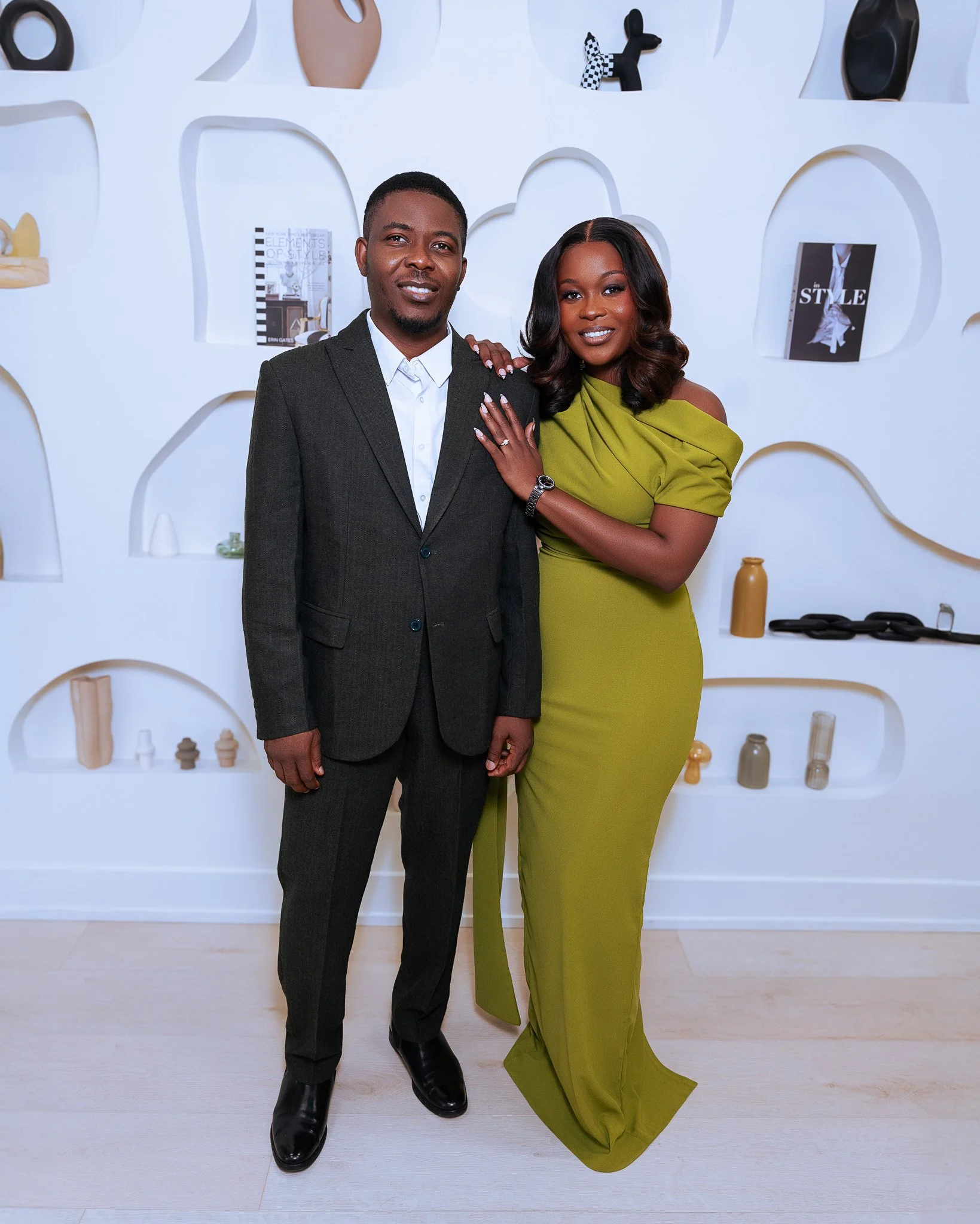 A man wearing a suit and a woman in a lime green dress pose together in front of a white wall with displays of decorative objects and books.
