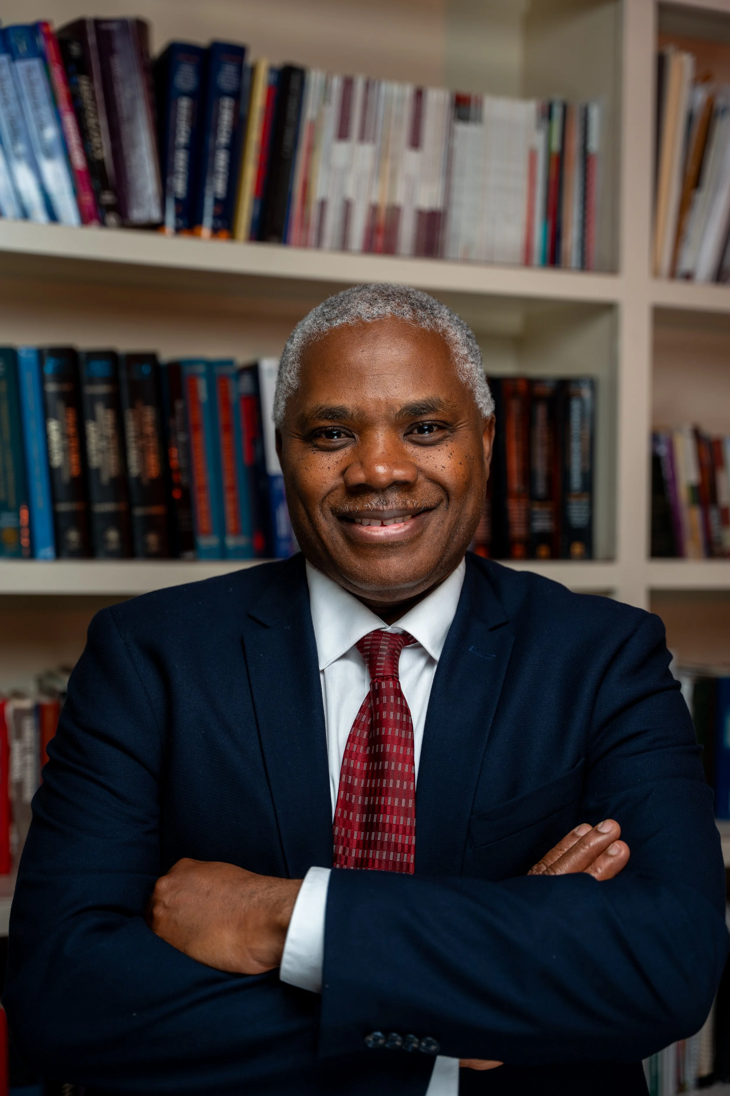 A smiling middle-aged man with gray hair, wearing a dark blue suit and red tie, sitting with arms crossed in front of a bookshelf filled with books.