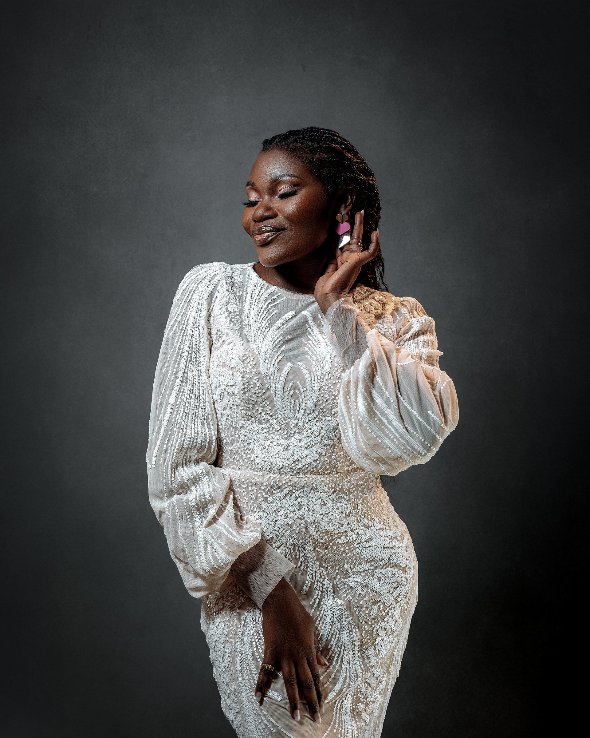 A woman with dark skin and braided hair wearing a white, textured gown with long sleeves, posing against a dark gray background, with her eyes closed and touching an earring.