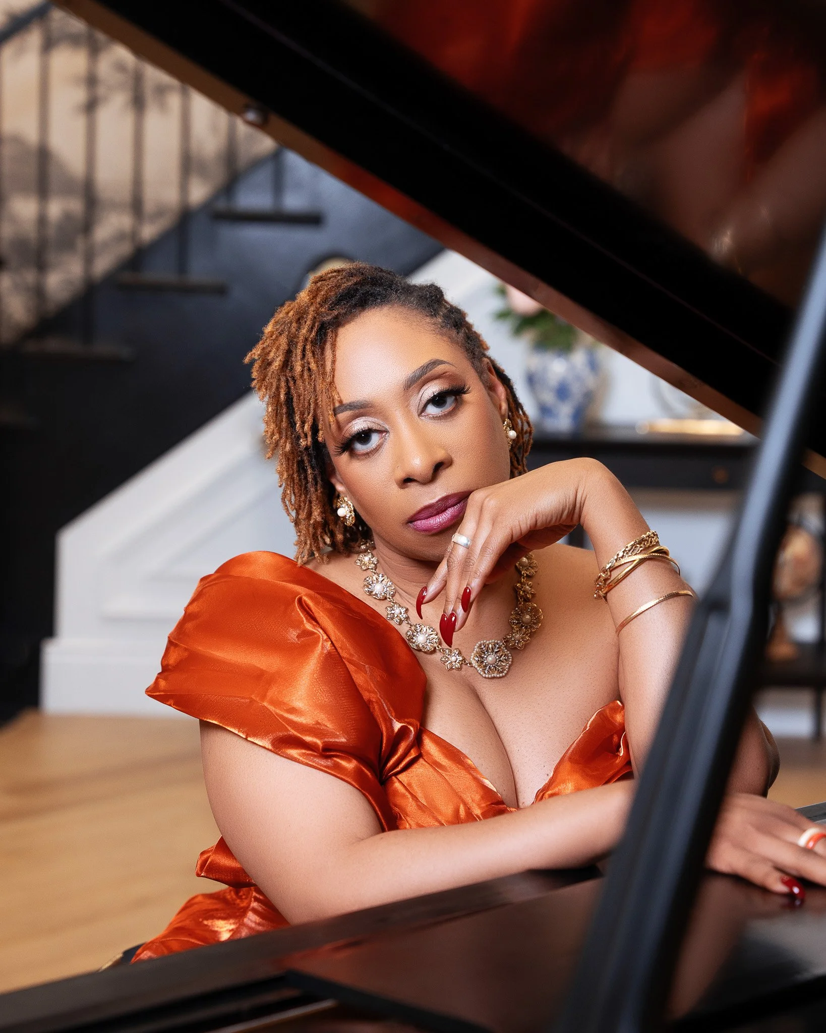 Close-up of a woman with curly reddish-brown hair wearing elegant jewelry and an orange satin dress, resting her chin on her hand while looking at a piano.