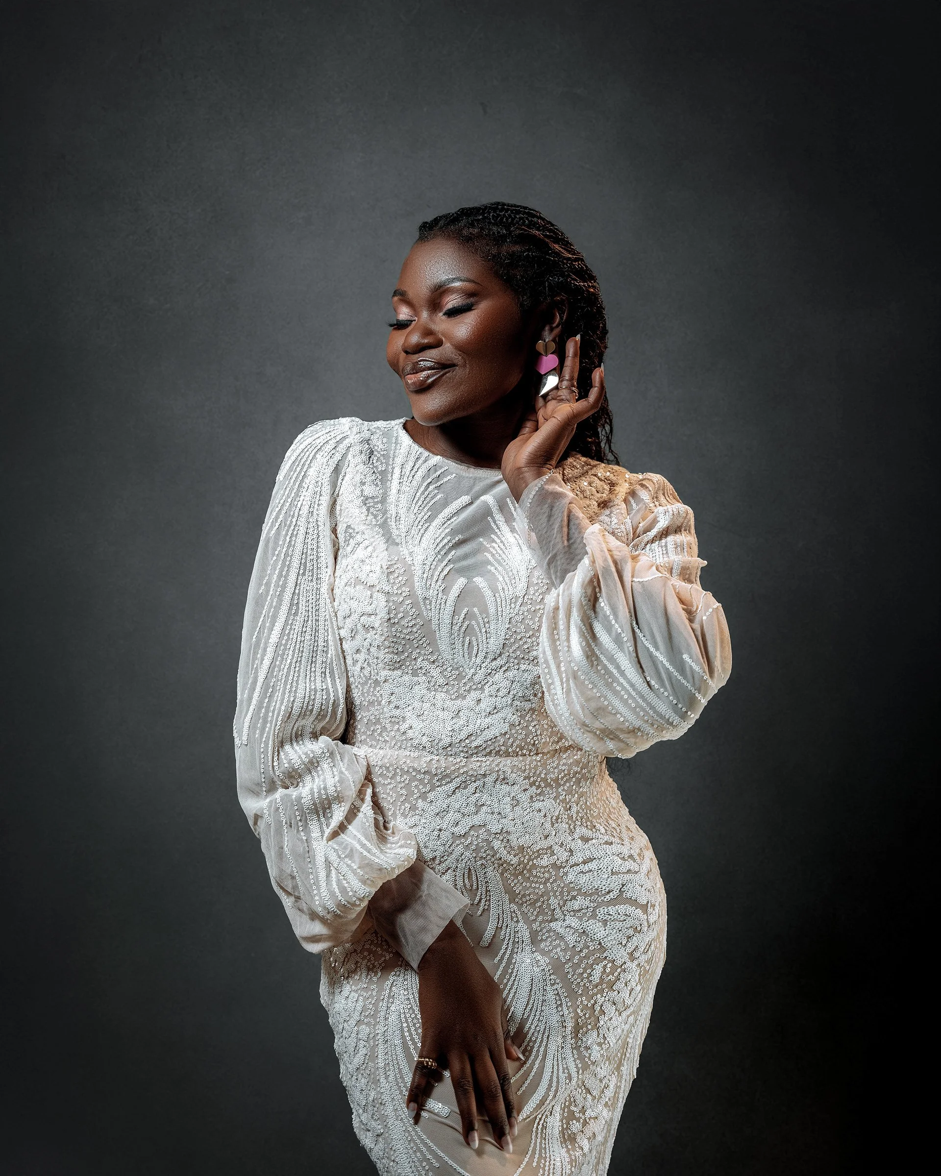 A woman with dark skin and long dreadlocks is wearing a white, intricately embroidered dress with long sleeves. She has her eyes closed and is smiling softly. She is touching her ear with her right hand, which is adorned with colorful earrings and ri