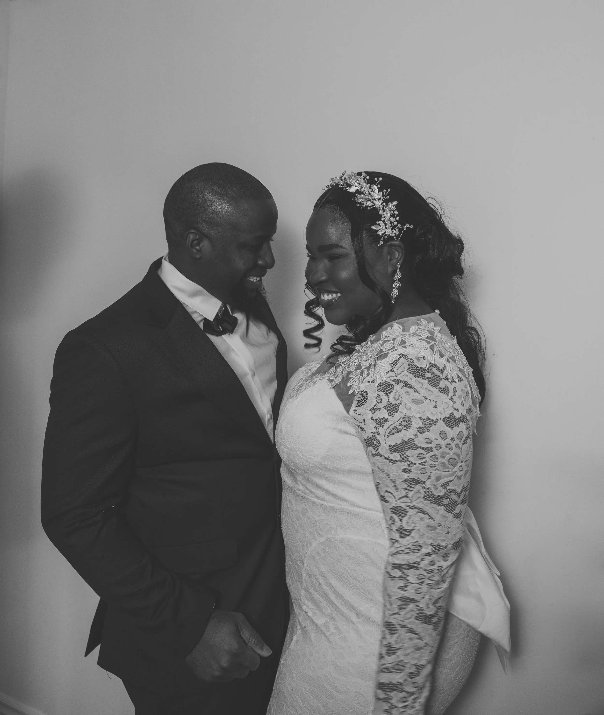 A black and white photo of a smiling couple, dressed in wedding attire, standing close with forehead to forehead, on a plain background.