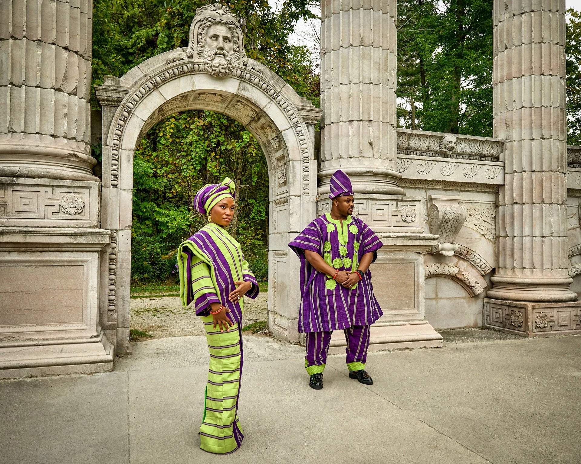Two people dressed in traditional African attire, standing in front of a historic stone gate with large columns and intricate carvings, surrounded by green trees.