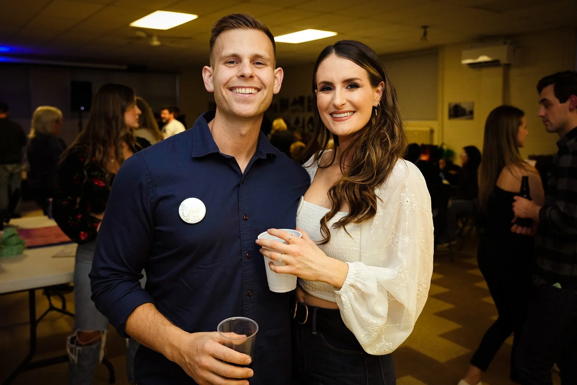 A man and woman smiling at a social gathering, the woman holding a cup, with other people in the background.