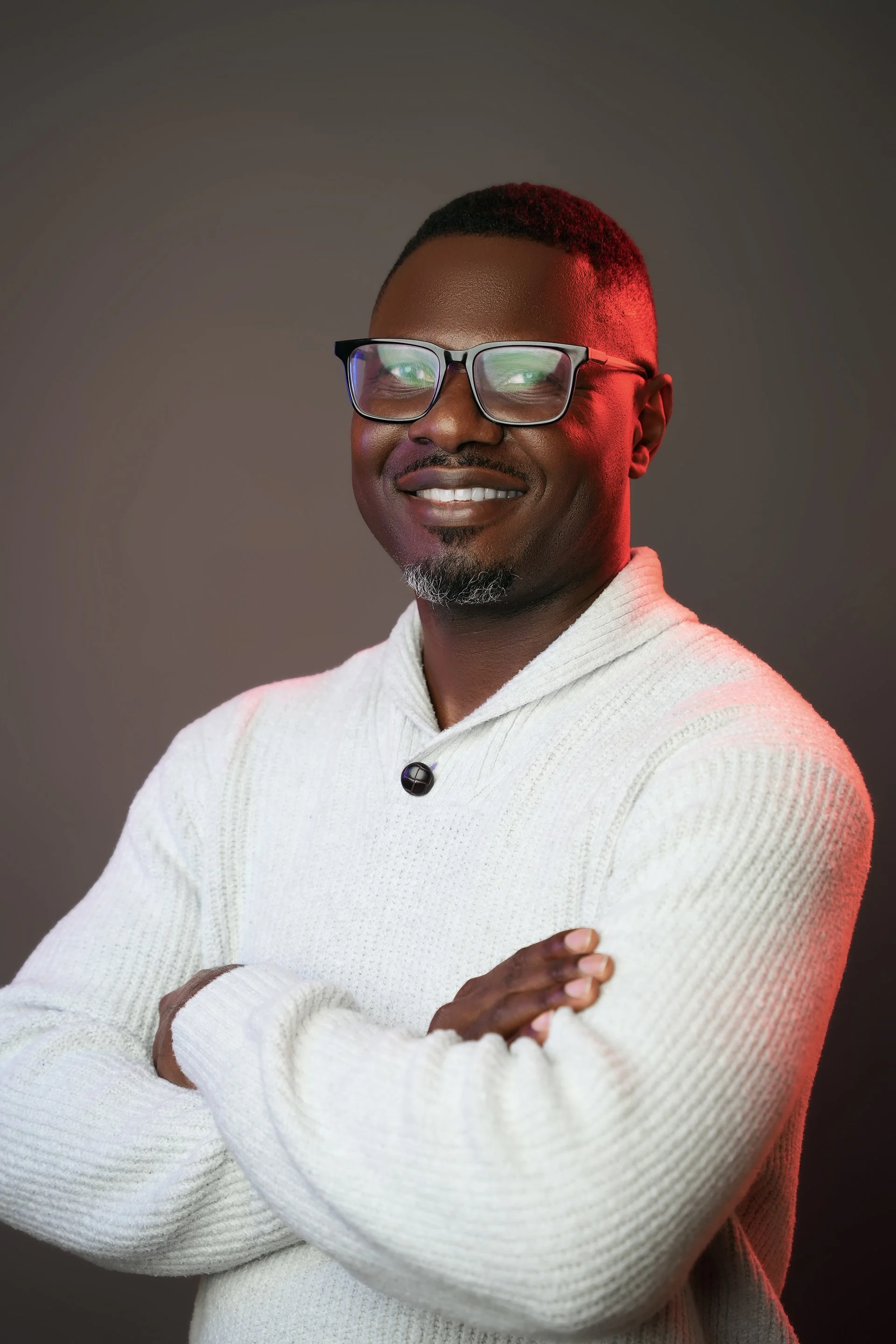 A smiling man with glasses, a goatee, and short hair, wearing a white sweater with his arms crossed, standing against a gray background with red and green lighting accents.