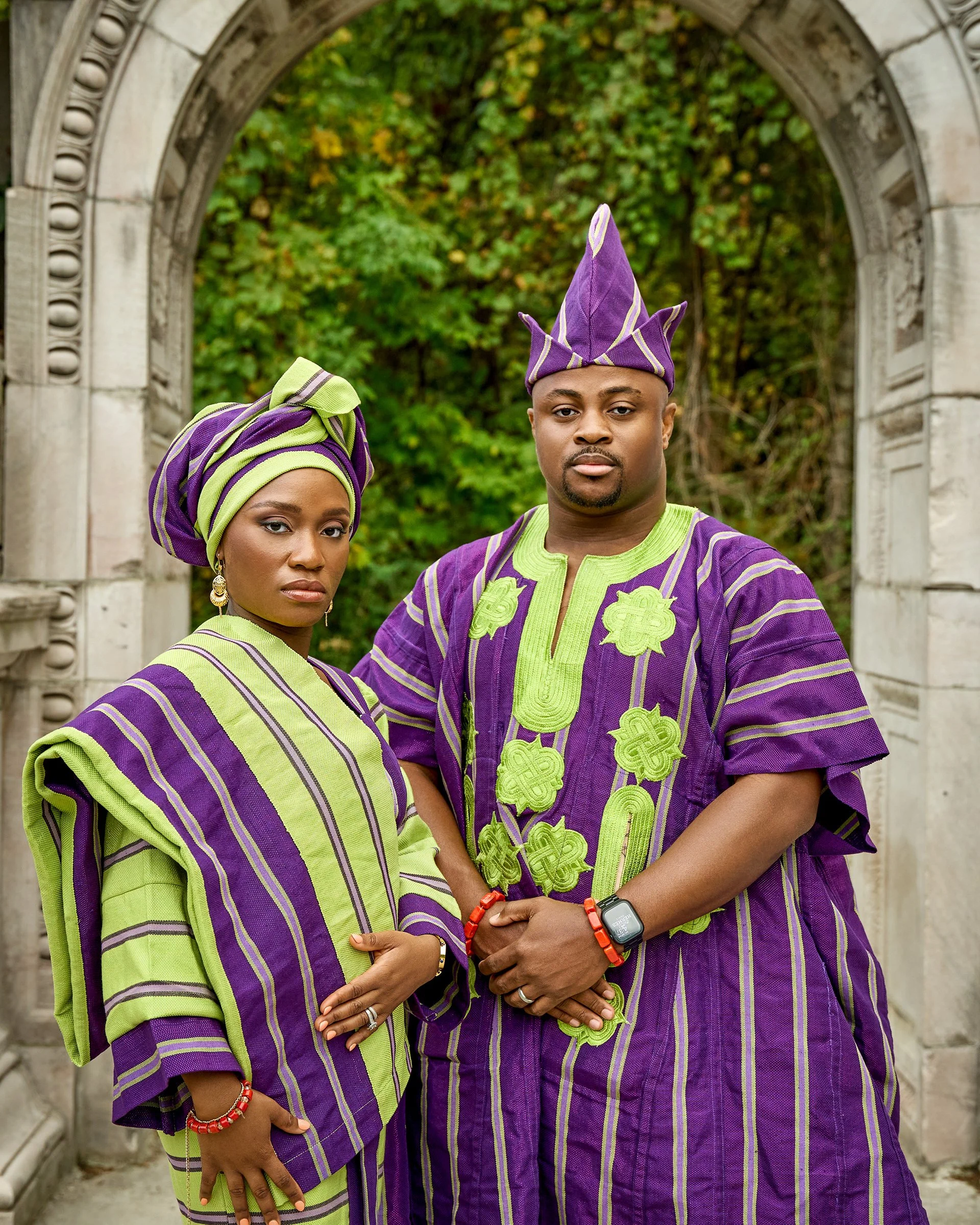 Two people, a woman and a man, dressed in matching purple and green traditional African attire, standing under a stone archway with a background of green trees.