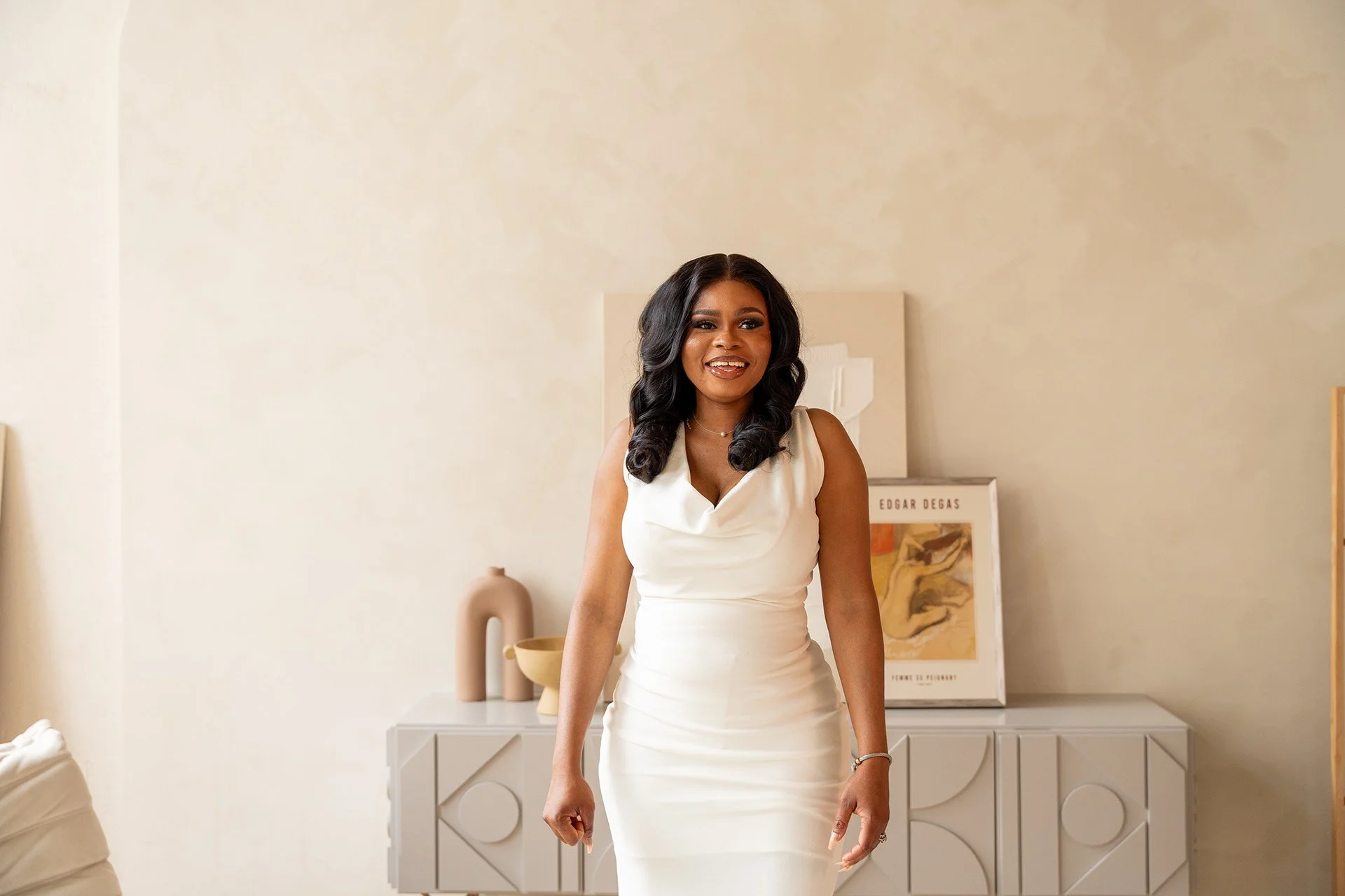 A woman with dark, voluminous hair styled in soft waves, wearing a sleeveless, fitted white dress with a draped neckline, standing in a neutral-toned living room. The background features minimalistic decor, including a framed piece of artwork, a beig