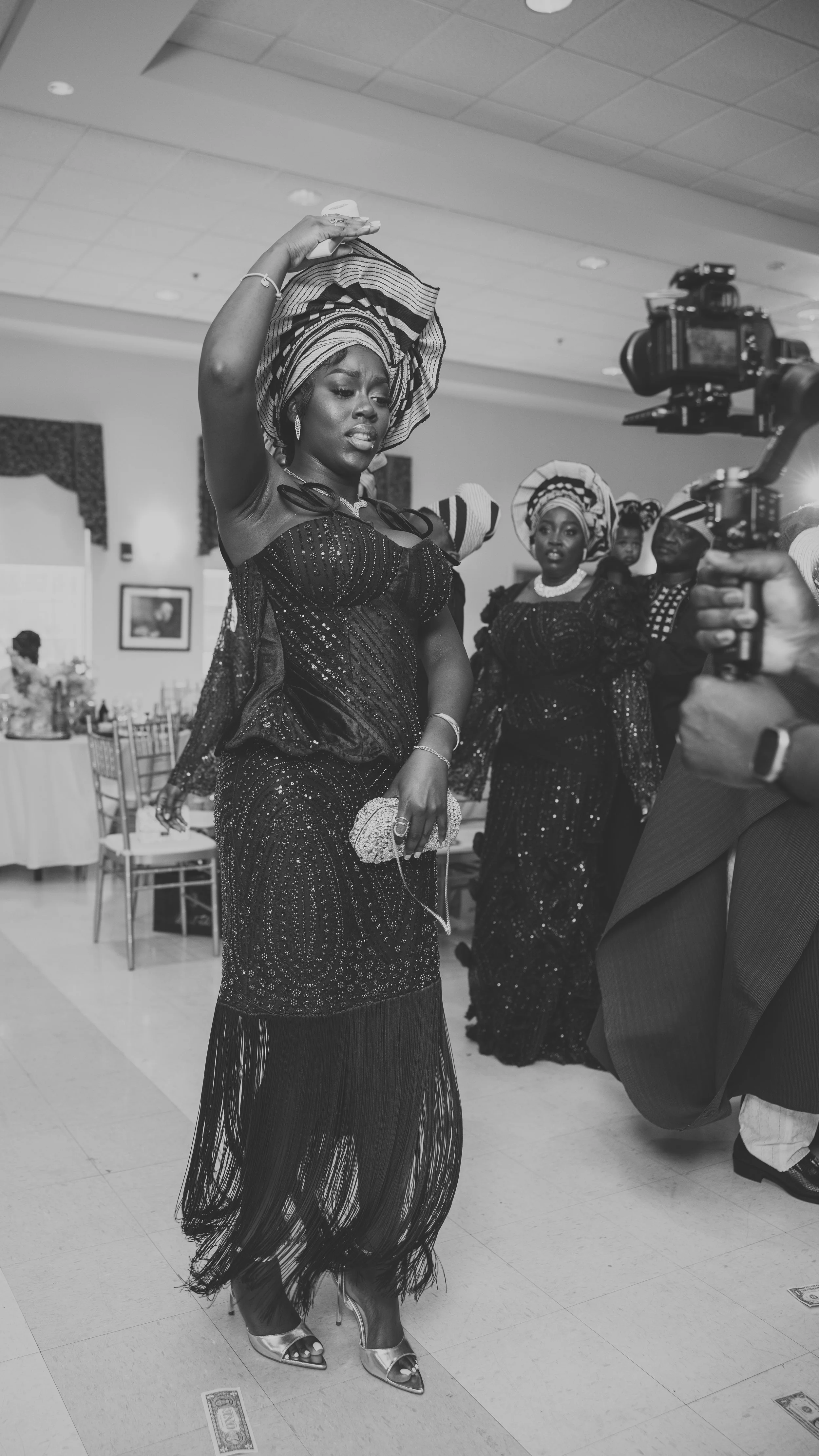 A woman dressed in an elegant black evening gown with sequins and fringes, wearing high heels, and a patterned headwrap, is dancing in a decorated indoor event space. She is holding a clutch and jewelry, with a serious expression. Several women in si