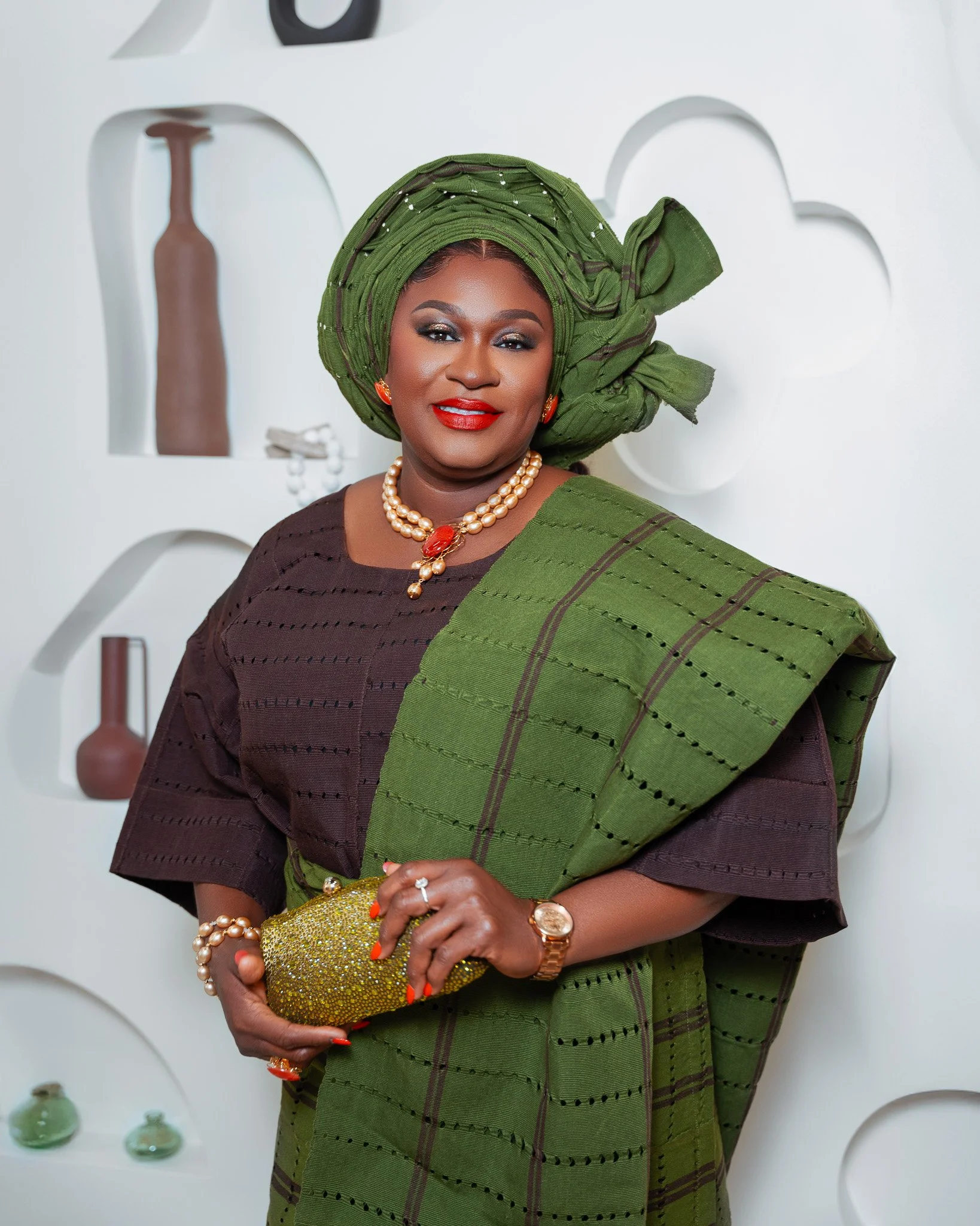 A woman dressed in traditional African attire, wearing a green headwrap and matching dress, with jewelry including a pearl necklace, earrings, and a watch, standing in front of a white decorative wall with open circle and bottle-shaped cutouts, holdi