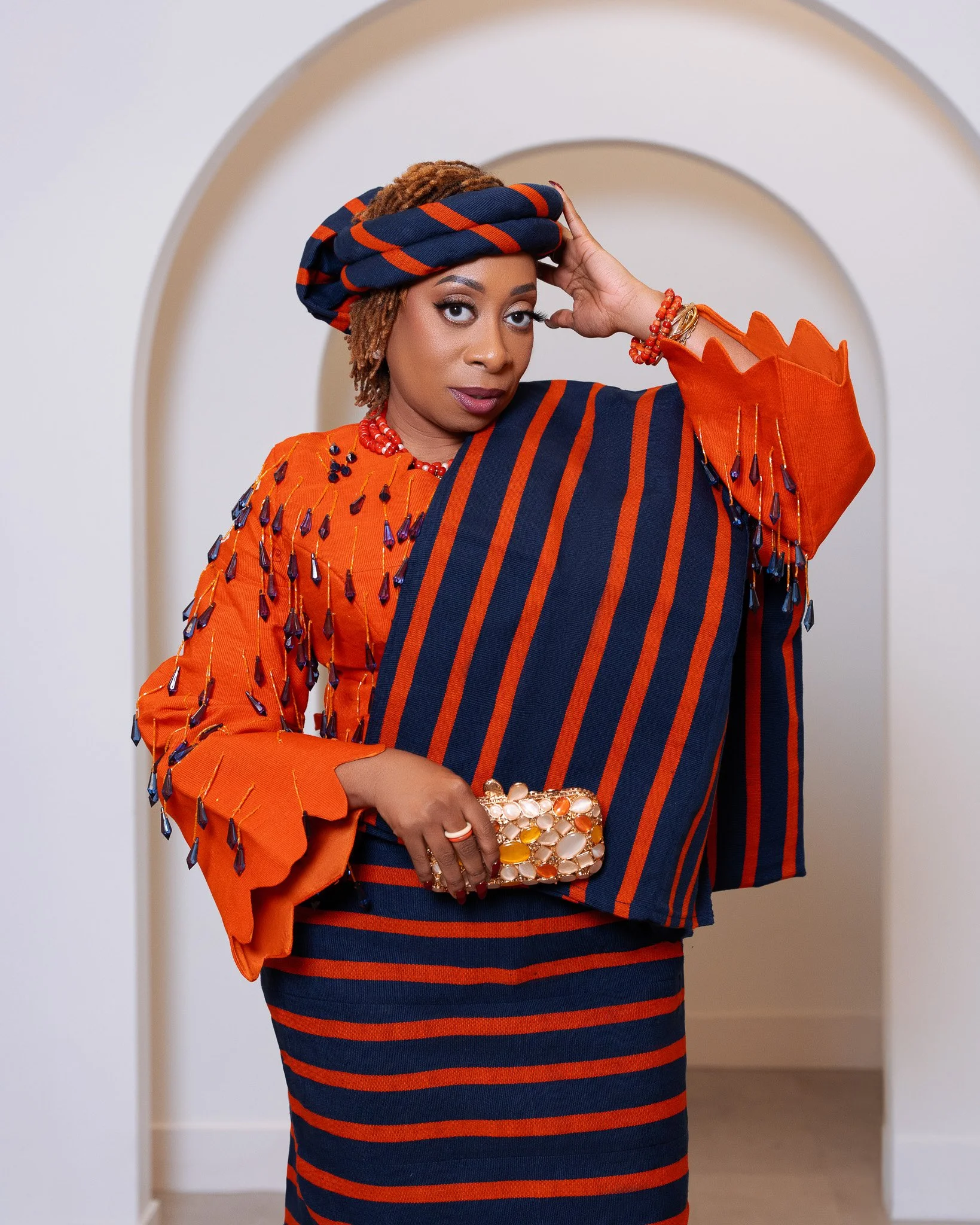A woman wearing a striped navy and orange dress with embellishments, an orange jacket with fringes and beads, a striped headwrap, and jewelry, holding a decorated clutch bag, posing in front of a white background with an archway.