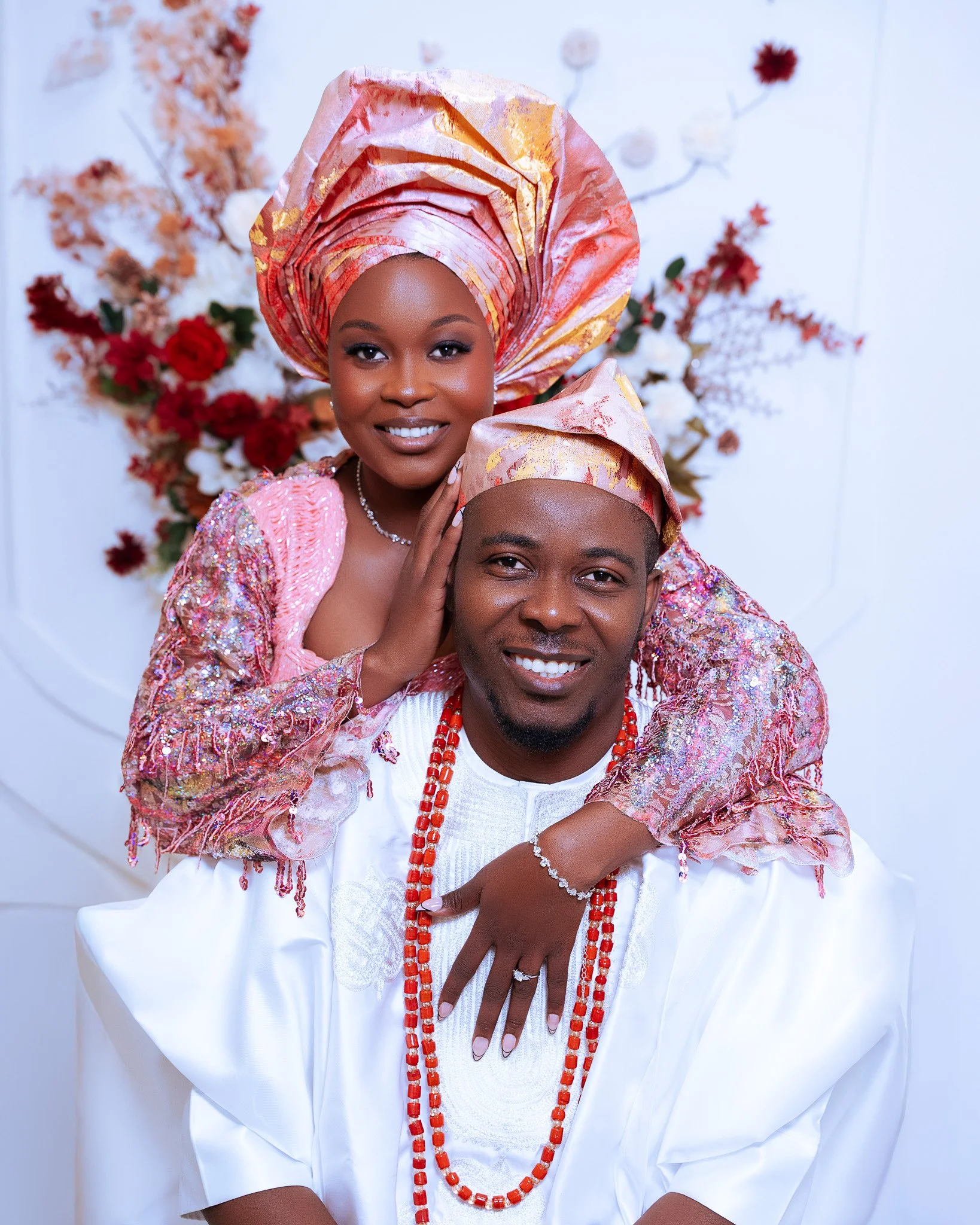 A woman wearing a colorful, patterned headwrap and matching attire, smiling next to a man in traditional white attire with a beaded necklace, in front of a backdrop of flowers and branches.
