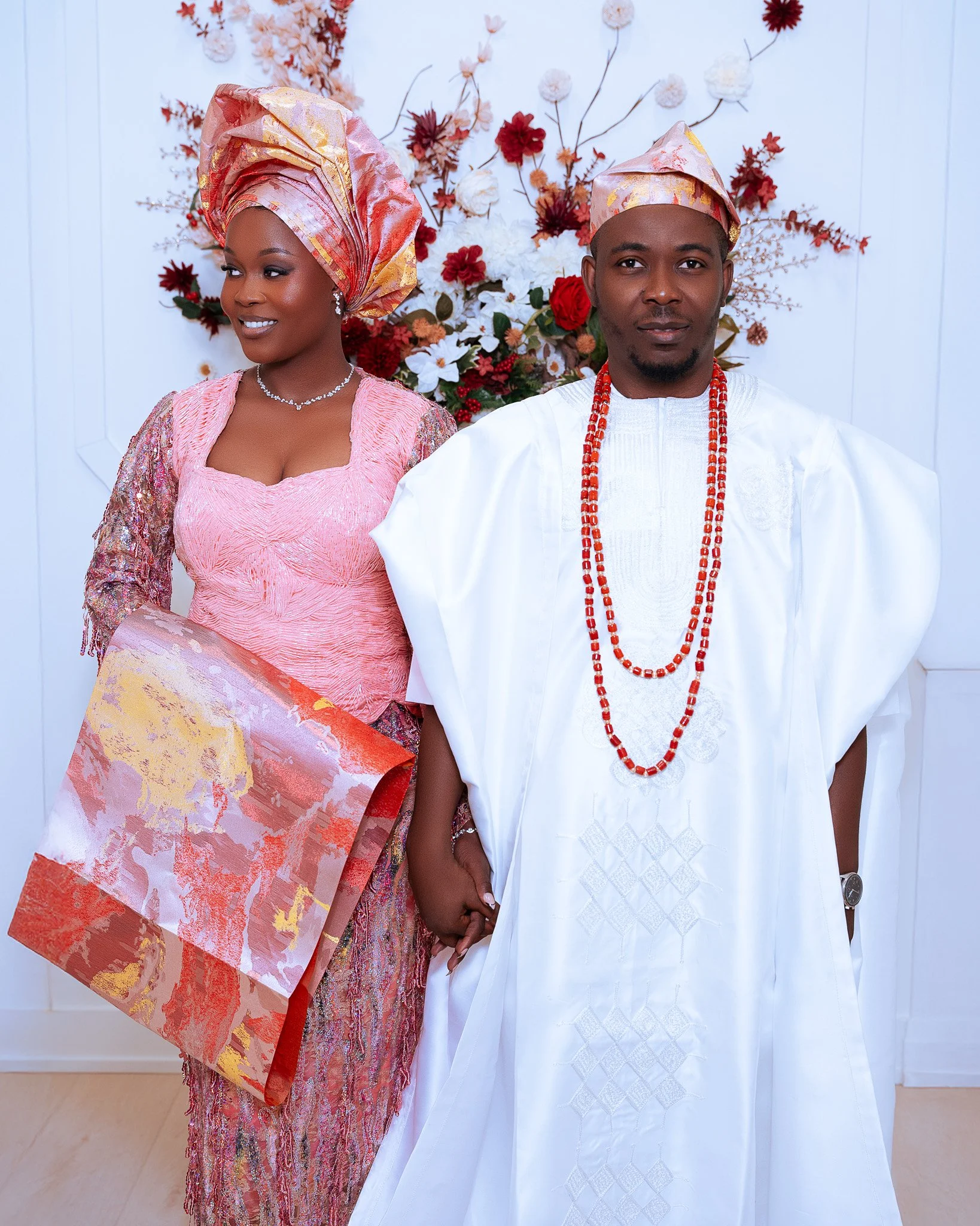 An African couple dressed in traditional attire, holding hands, standing in front of a floral backdrop, celebrating a cultural event or wedding.