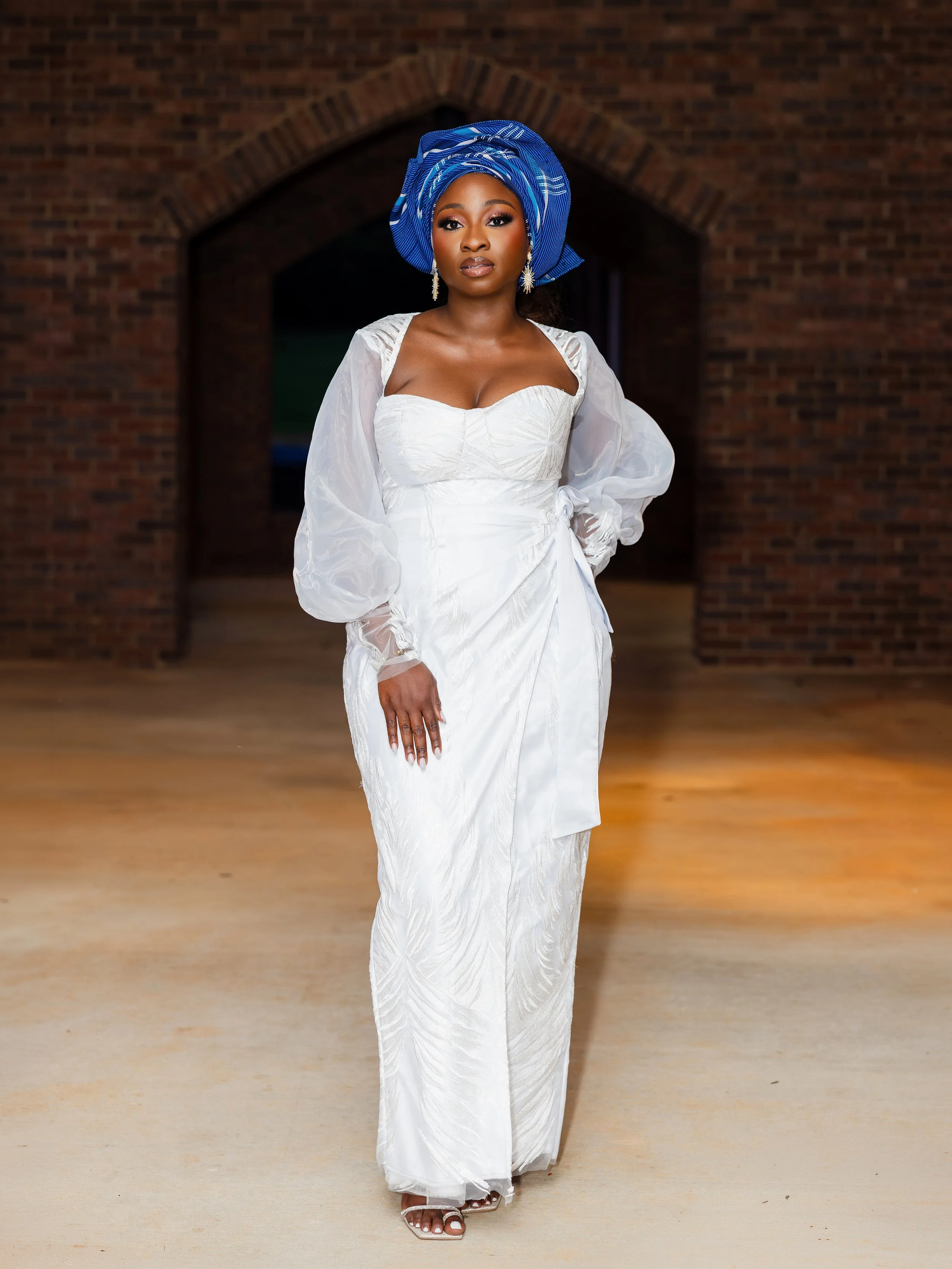 A woman wearing a white dress with puffy long sleeves and a blue headwrap, standing in front of a brick archway. She has earrings and makeup, and is standing on a wooden floor.