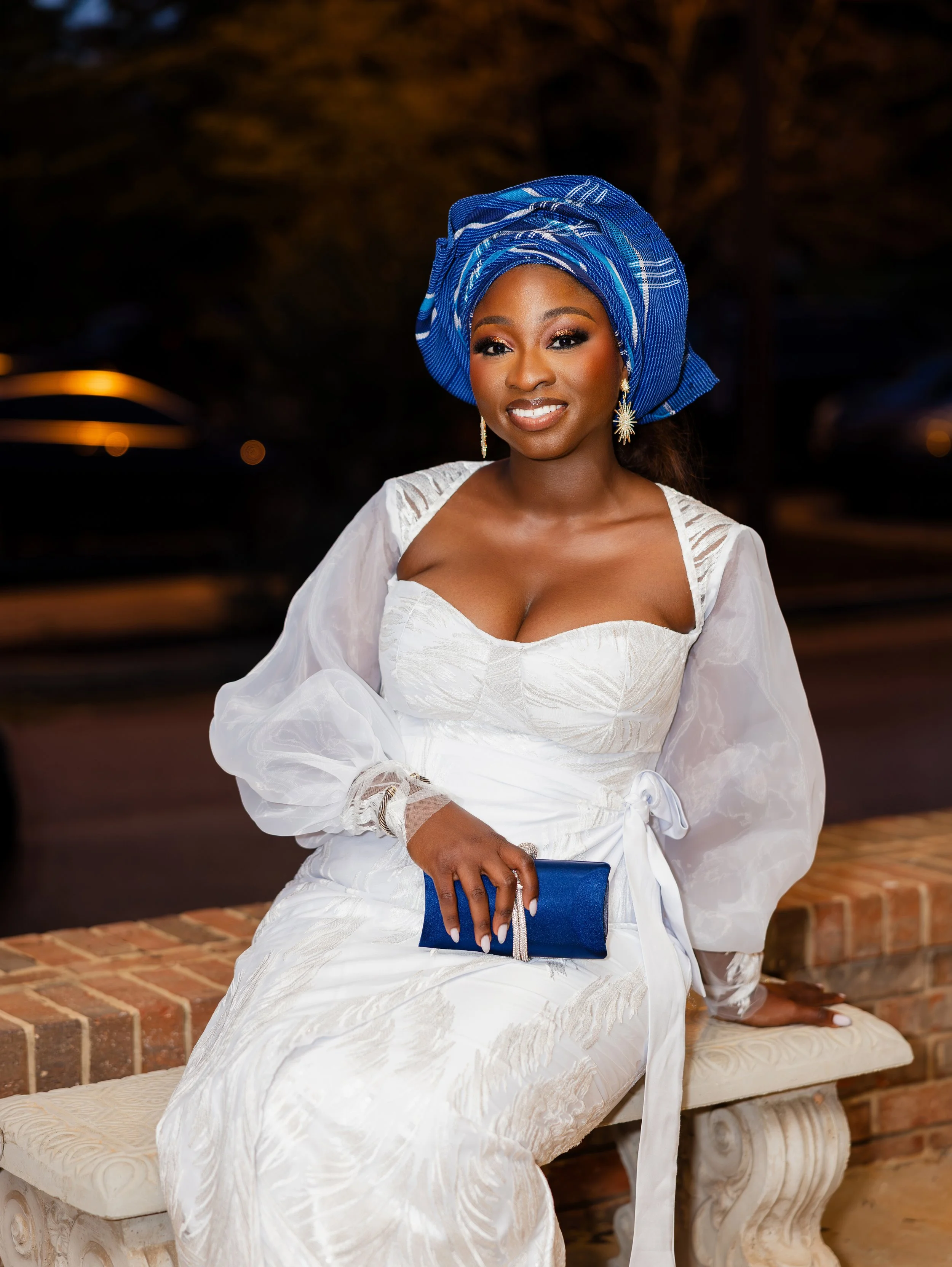 A woman sitting on a stone bench outdoors at night, wearing a white dress and a vibrant blue headwrap, holding a matching blue clutch, with a warm smile.