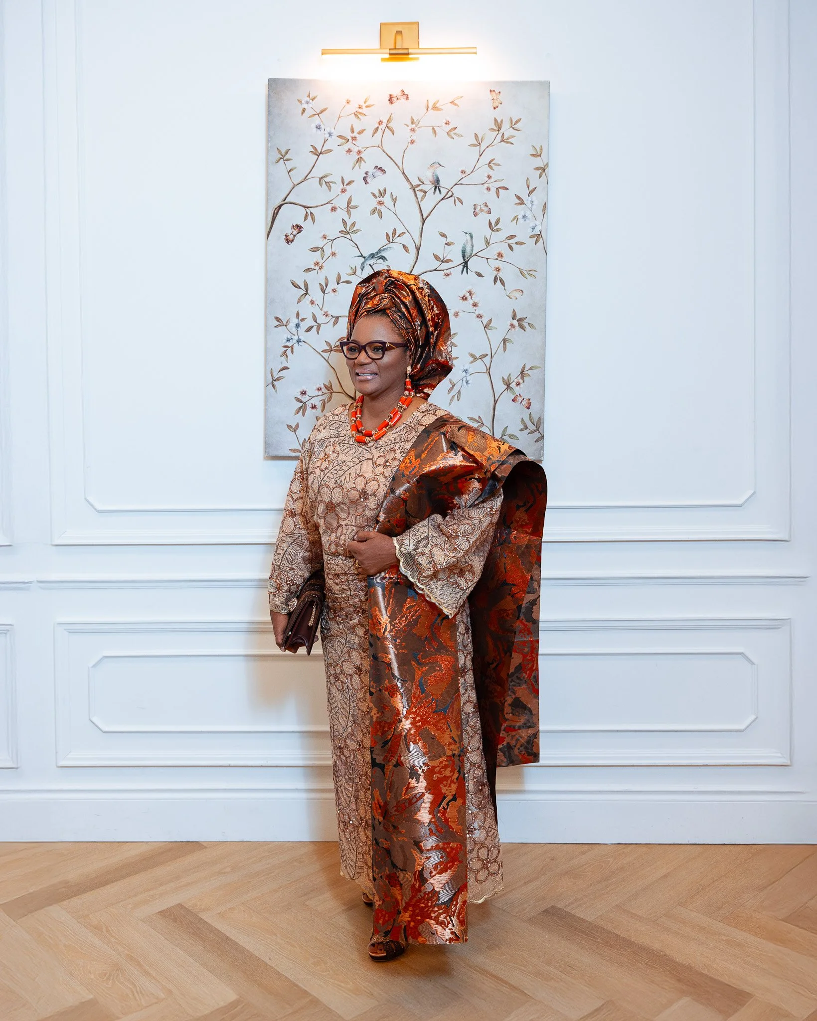 A woman dressed in traditional African attire standing in front of a wall with a framed artwork of birds, butterflies, and flowers. She is wearing glasses, a headwrap, a necklace, and holding a handbag.